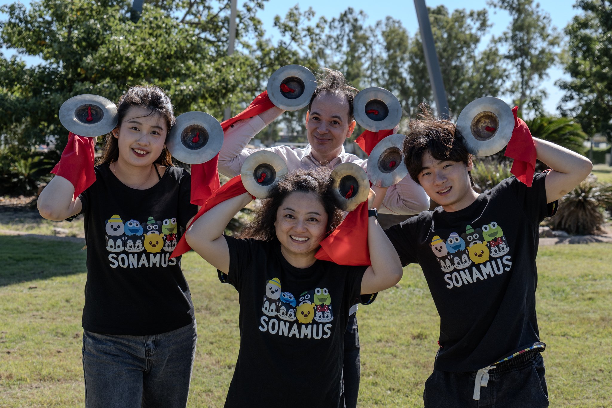 Four people outdoors holding circular metal objects above their heads, smiling. They are wearing black T-shirts with cartoon characters and the text "SONAMUS". The background shows trees and a clear sky.