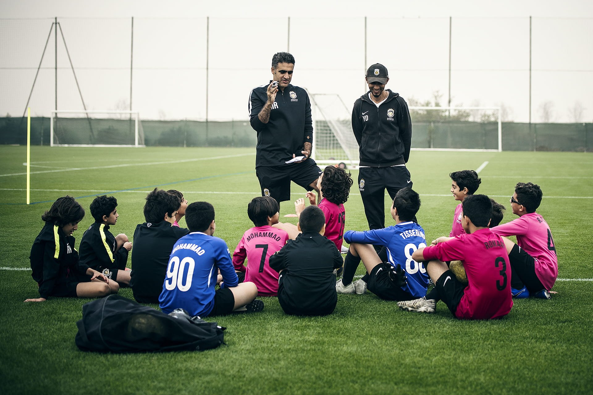 Soccer coach talking to children during a training session on a soccer field, with kids sitting on the grass, some wearing pink and blue uniforms.