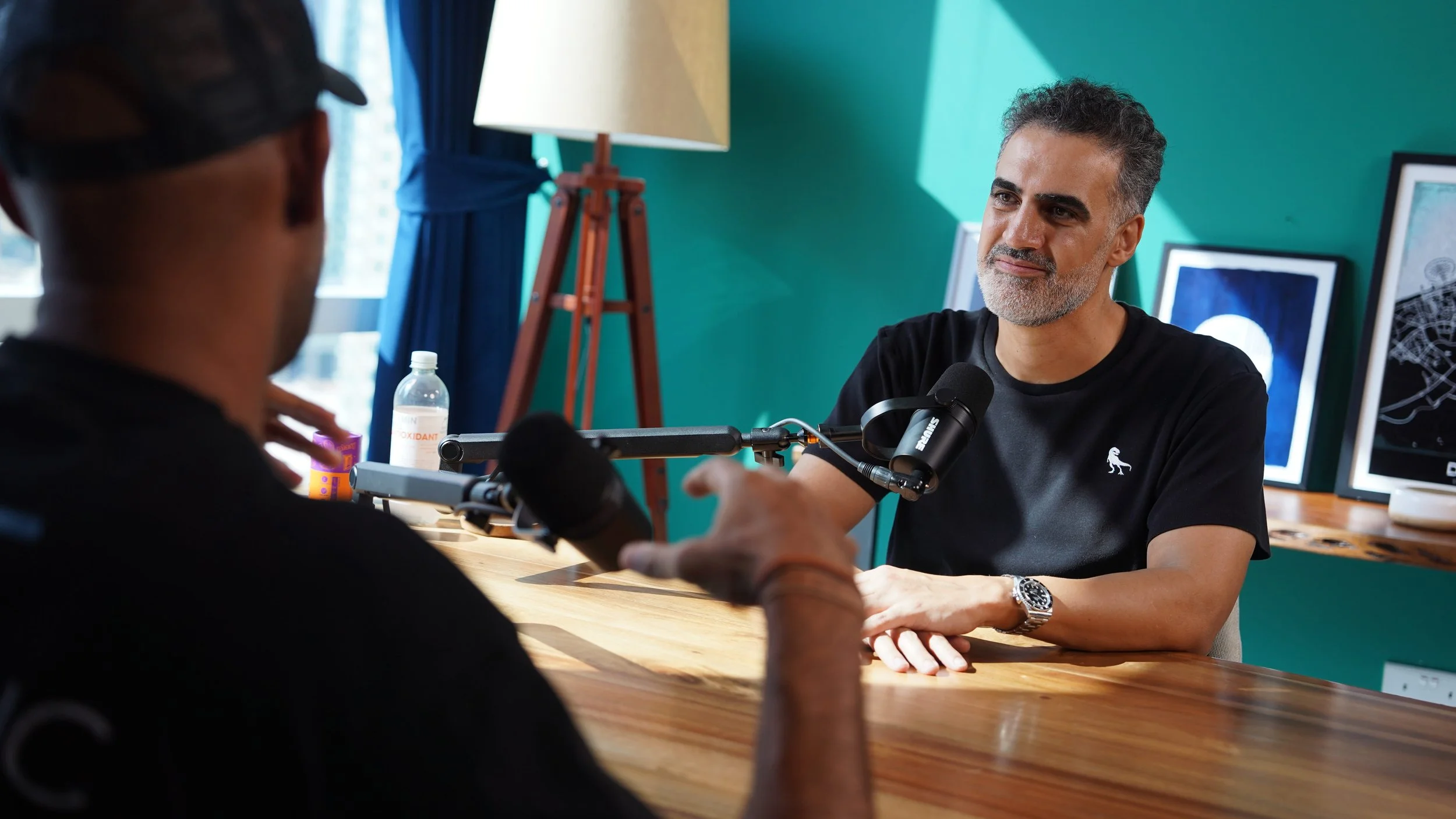 Two men having a conversation in an indoor setting, with one man facing the camera and the other with his back to the camera, seated at a wooden table. There are framed pictures and a lamp in the background. The man facing the camera has graying hair and is wearing a black shirt.