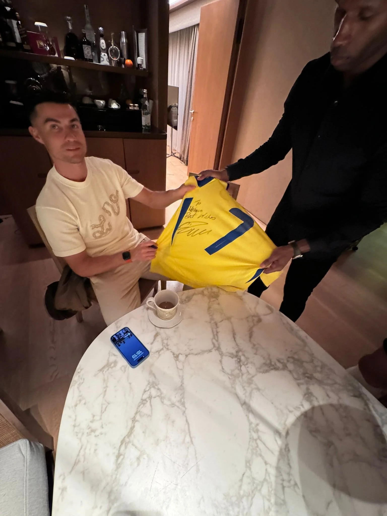 A man sitting at a marble table holding a yellow bag with two people standing nearby. A mug of tea and a blue phone are on the table. The background features a wooden cabinet with bottles and other items.