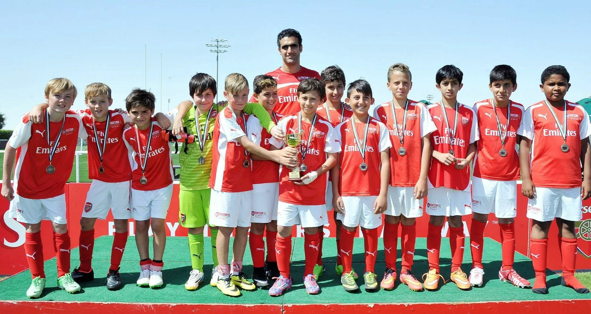 Youth soccer team in red and white uniforms celebrating with coach on a field, holding a trophy and medals.