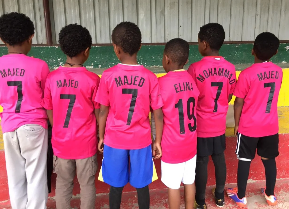 Six young children standing side by side, wearing pink sports jerseys with their names and numbers on the back, facing away from the camera.