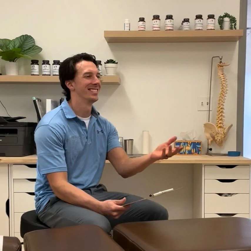 A man in a light blue polo shirt sitting on a stool in a medical office, smiling and gesturing, with shelves of supplements, a spine model, and office supplies in the background.