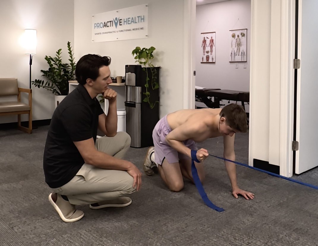 A chiropractor is observing a young man doing resistance band exercise on the floor inside a clinic.