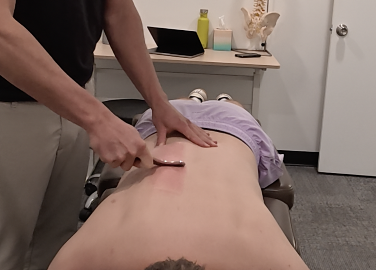 A person lying face down on a massage table receiving a massage or therapy, with a therapist applying pressure with a tool on their back in a clinical setting.