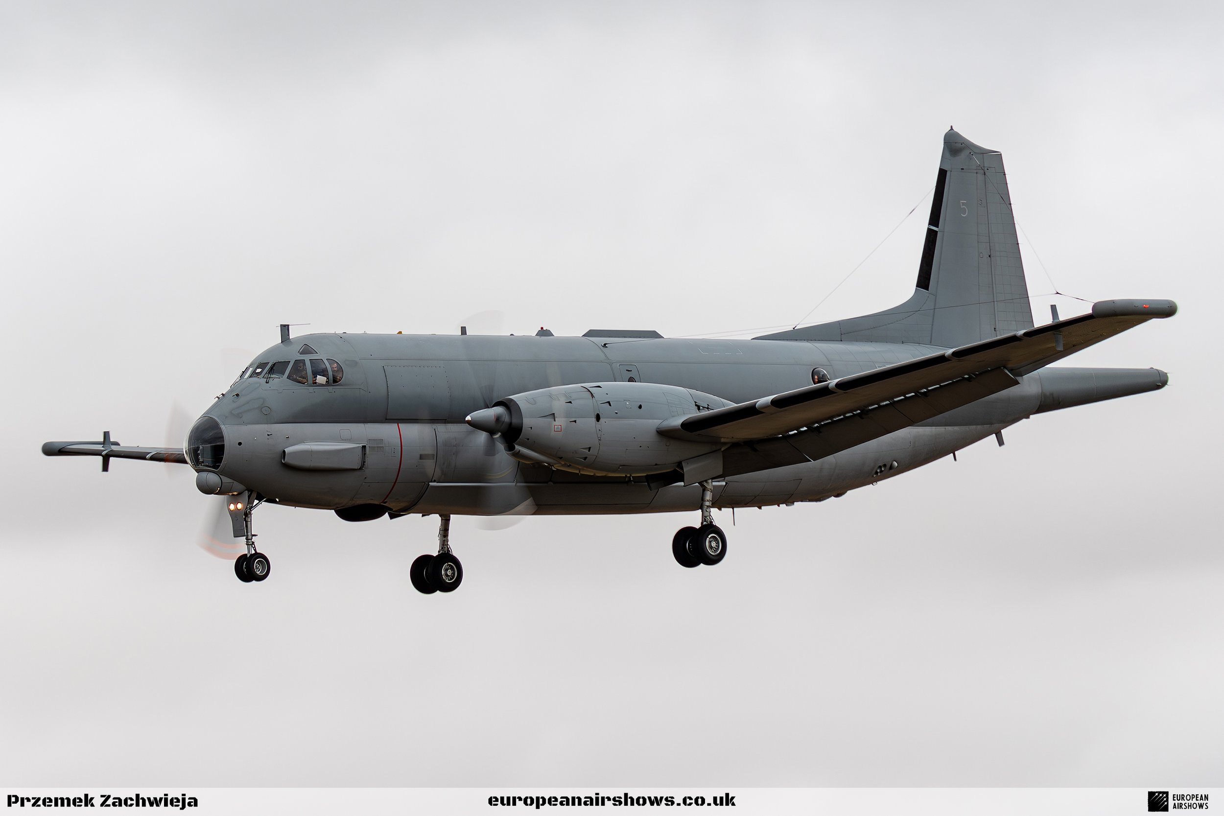 A gray military cargo aircraft flying in an overcast sky. The aircraft has two engines mounted under the wings, a high-mounted tail, and tricycle landing gear.