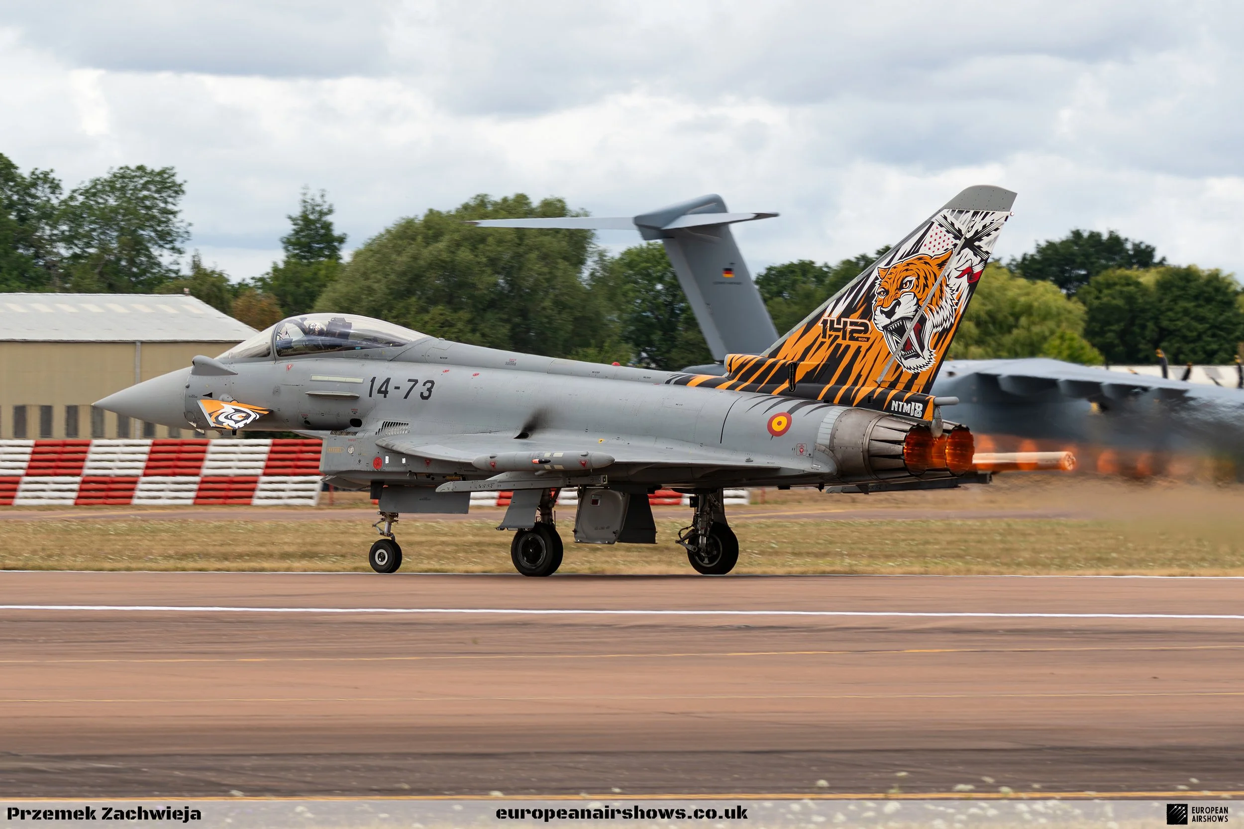 A military fighter jet taking off with afterburners on, on a runway with trees and hangars in the background. The jet has a tiger design on the tail.