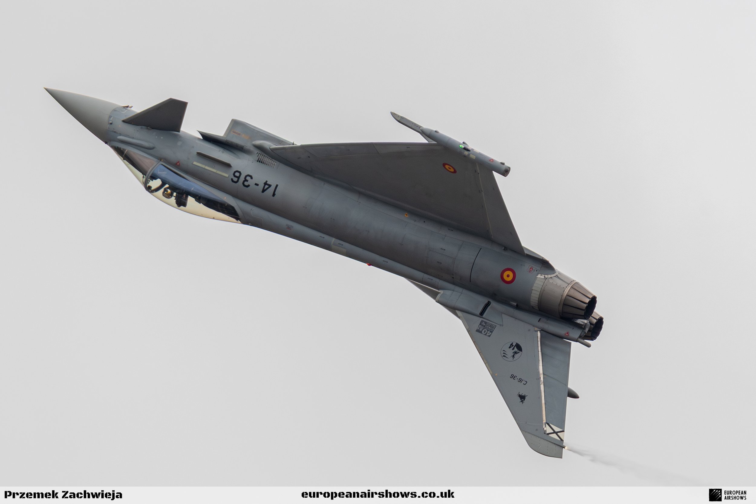 A military jet airplane flying in the sky, angled upward with vapor and condensation around the wings, against a gray overcast background.