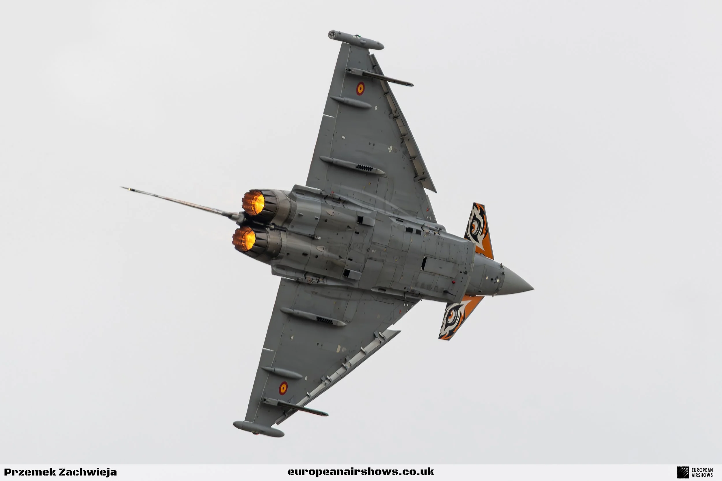 A fighter jet aircraft with afterburner flames, flying through a cloudy sky, with logo and markings on its tail fin.