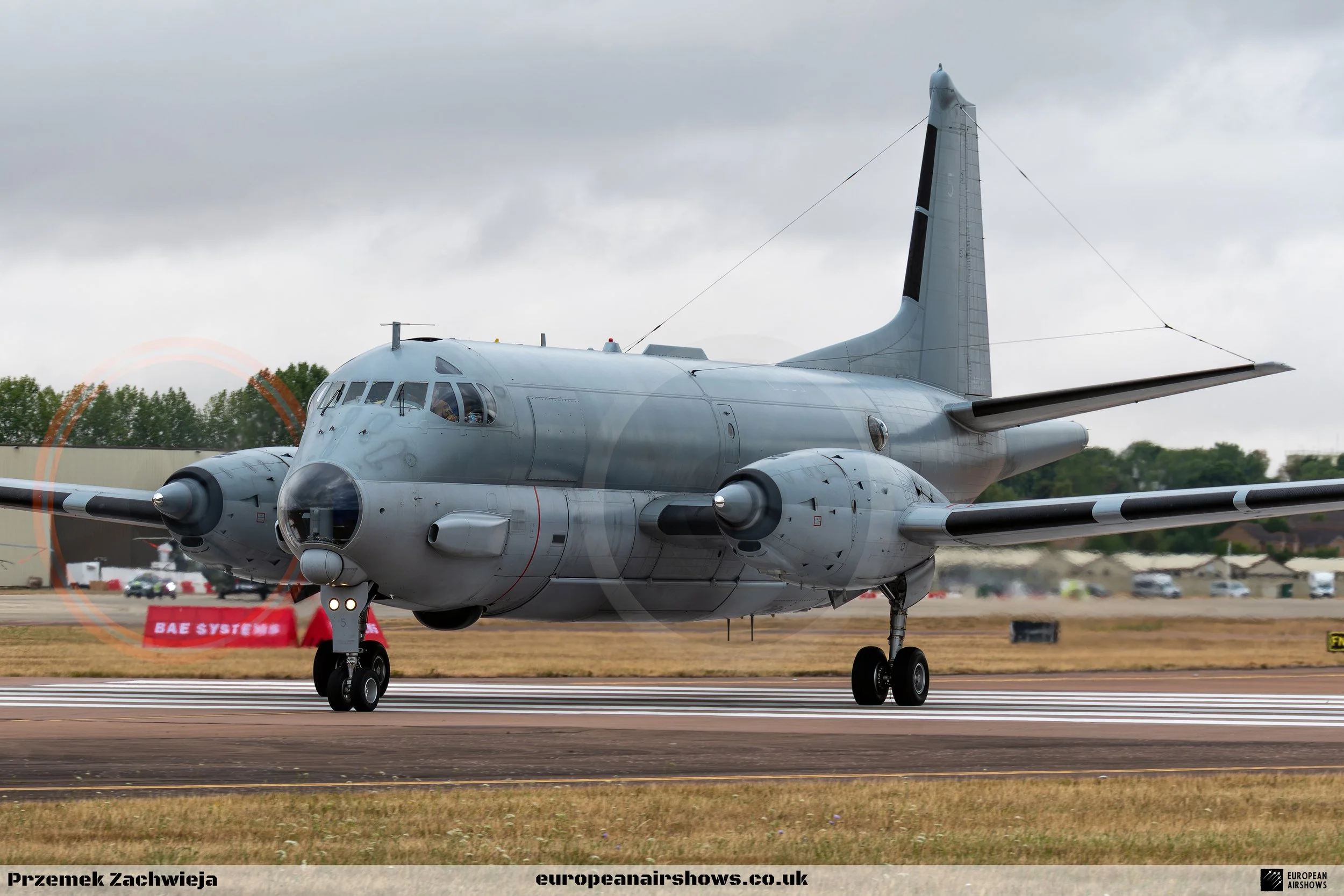 A military aircraft on the runway, painted in a flat gray color, with jet engines on each wing, a high tail fin, and a cockpit with several windows. The background has trees and a cloudy sky.