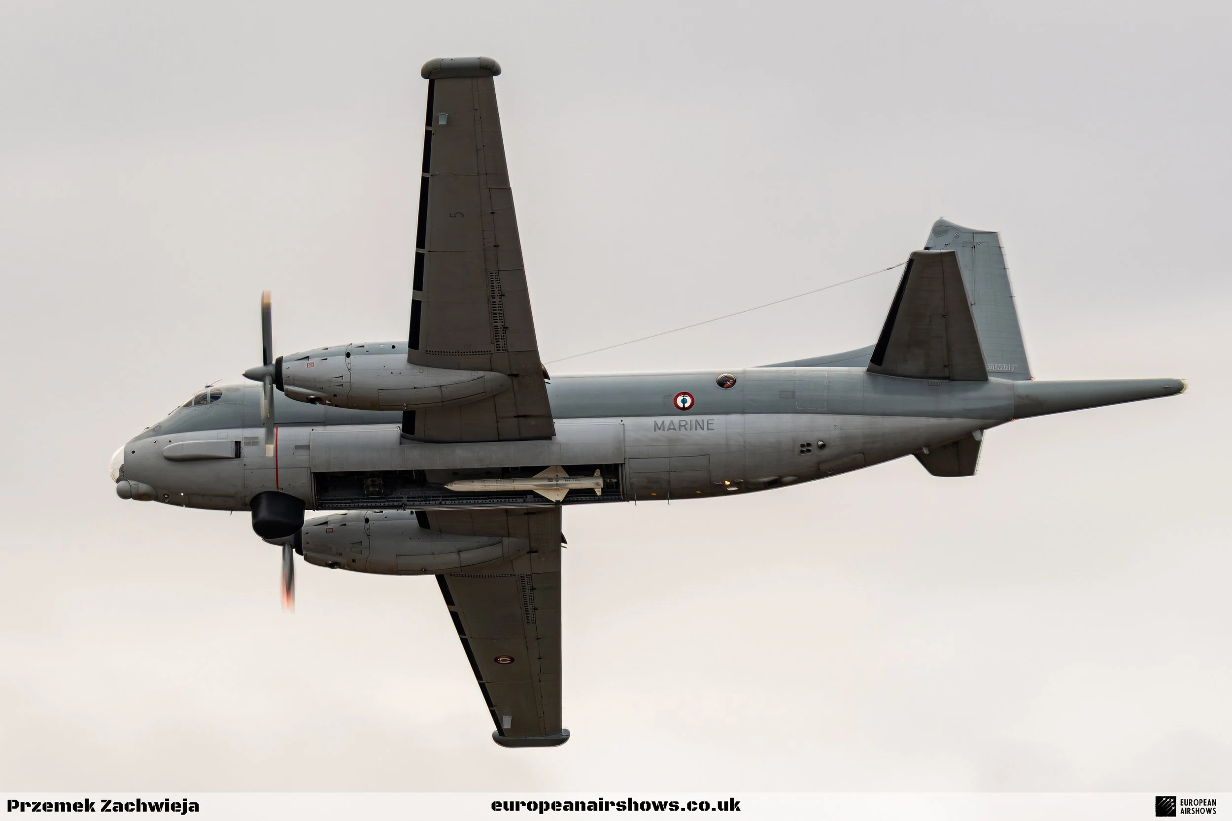 A gray military aircraft flying in the sky, with some clouds in the background, and the aircraft has the word 'MARINE' written on its side.