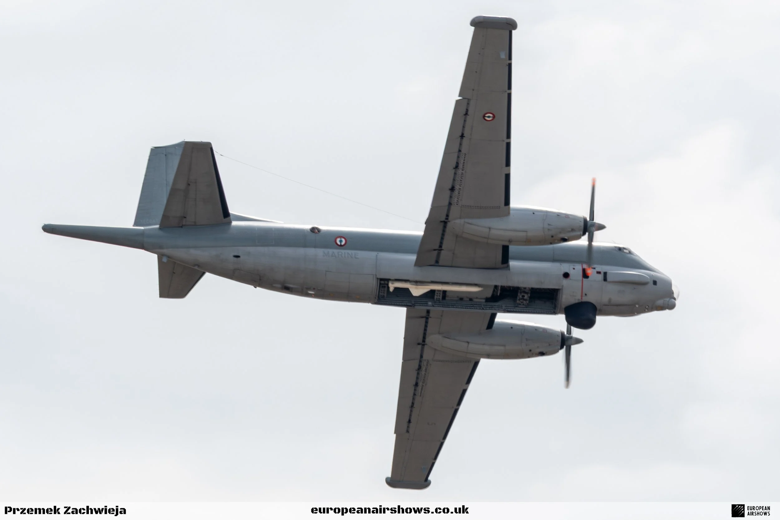 A military aircraft flying in the sky, viewed from below, with gray clouds in the background.