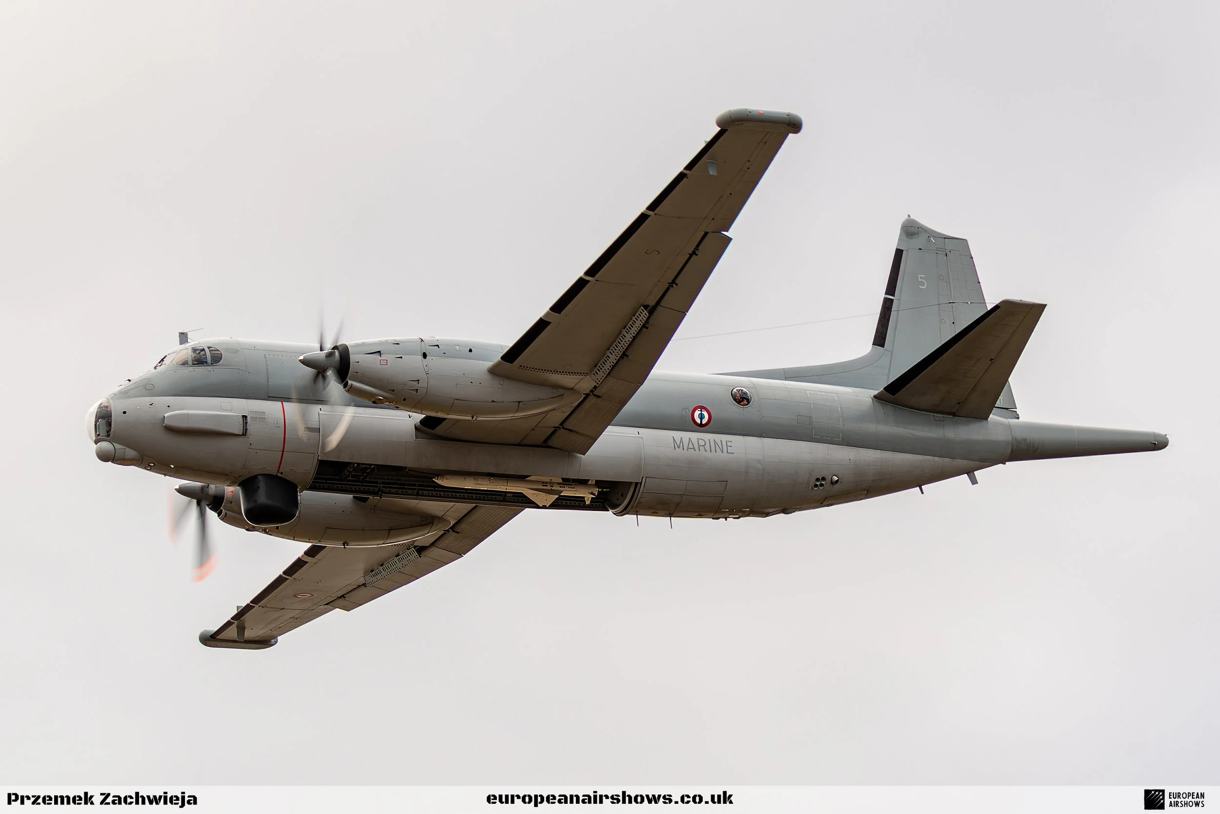 A gray military aircraft in flight with the word 'MARINE' on its side and a circular emblem near the cockpit, set against a cloudy sky.