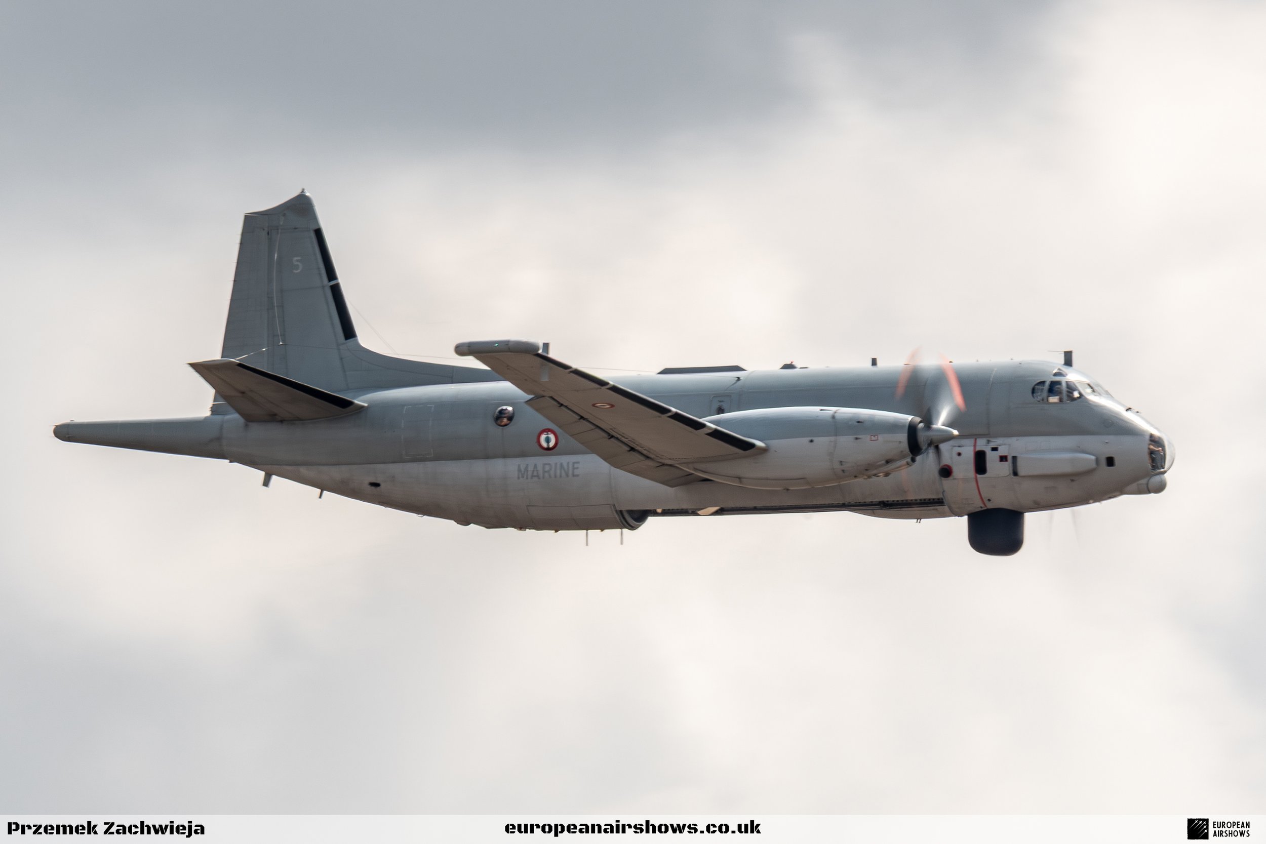 A military aircraft in flight against a cloudy sky, with a propeller at the nose and featuring a gray camouflage pattern.