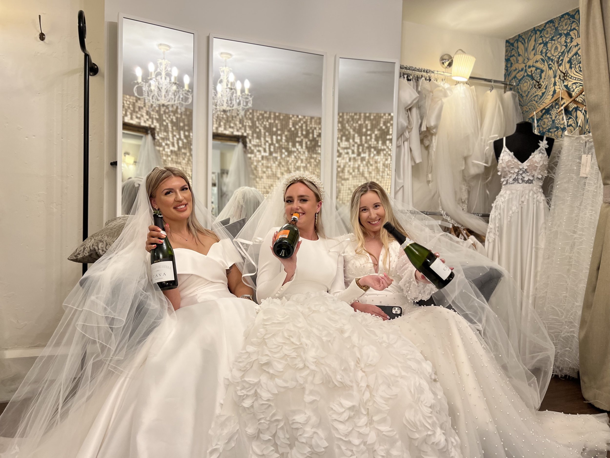 Three brides in couture bridal gowns smiling to the camera whilst sitting on a sofa and drinking cava from the bottle