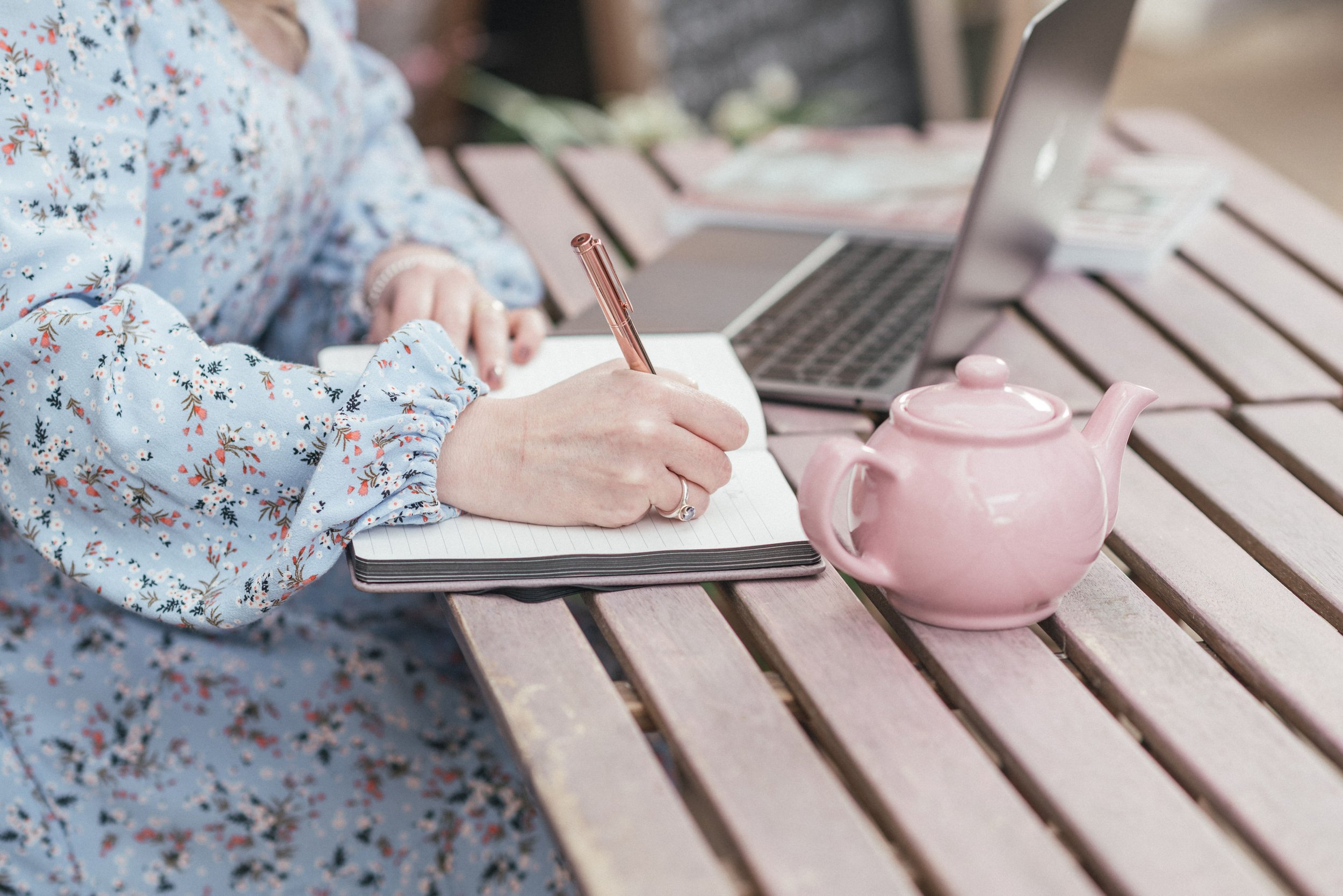 Woman journaling with a pot of tea, notebook and laptop