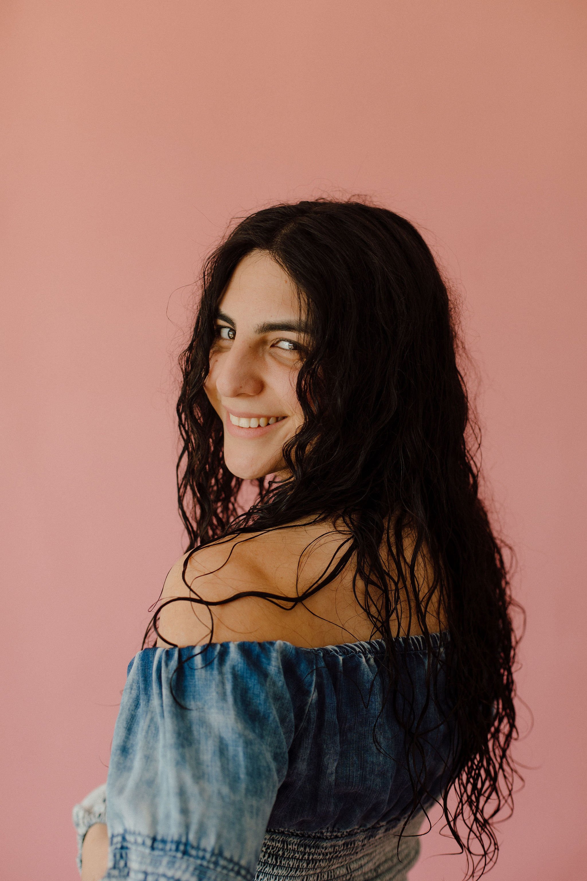 Woman with long curly black hair smiling over pink background wearing an off-the-shoulder denim top.