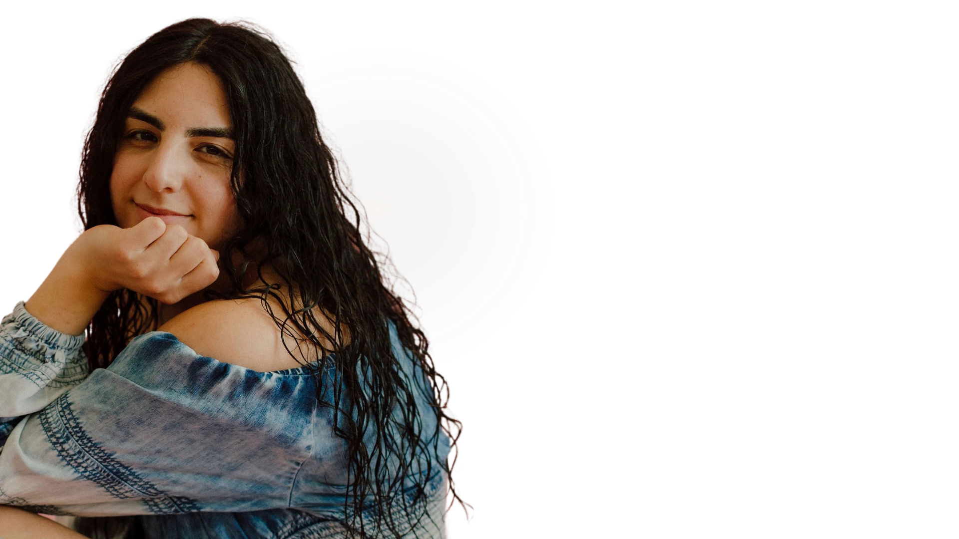 A young woman with long dark curly hair and a slight smile, wearing a blue patterned off-shoulder top, resting her chin on her hand.