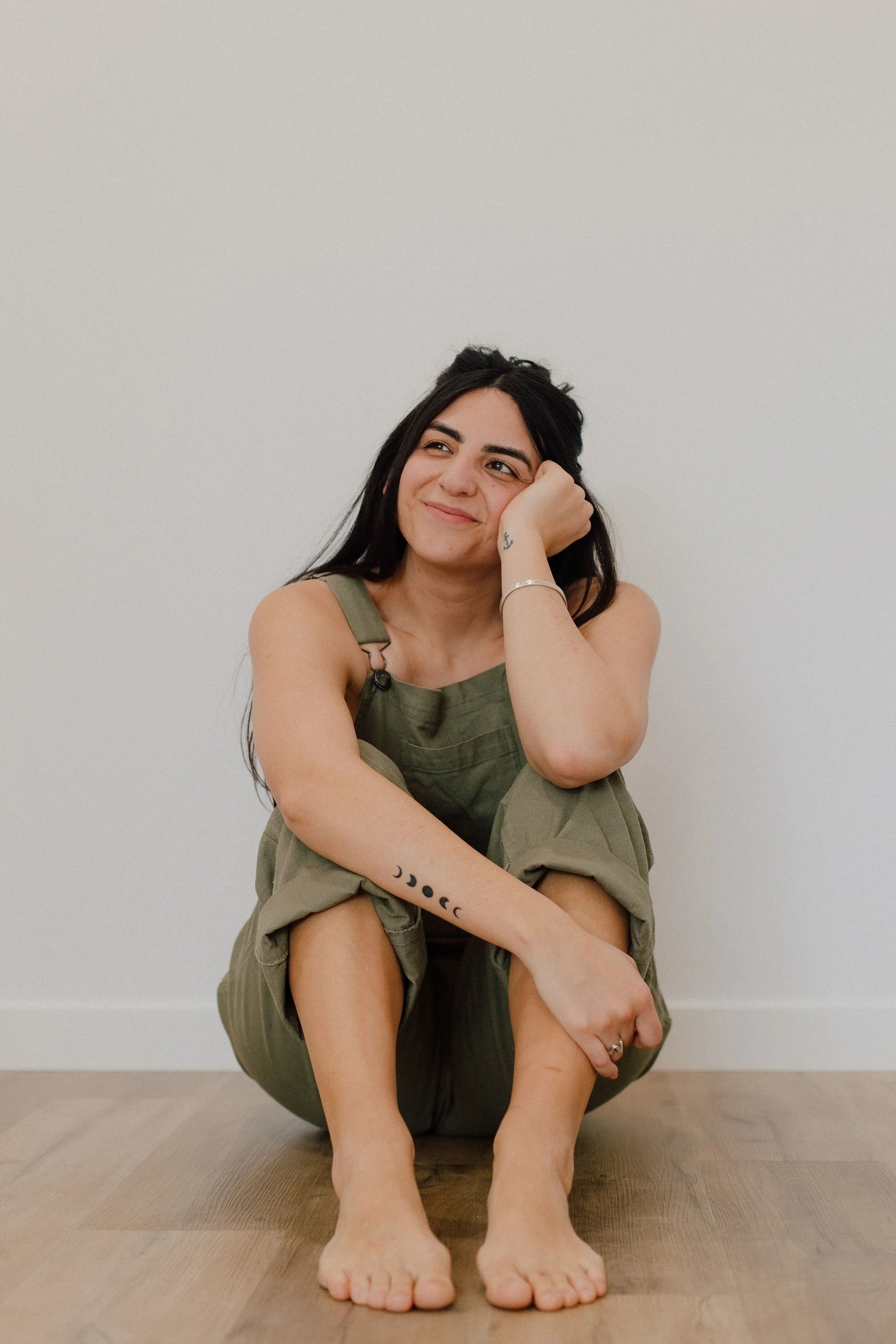 A woman with long dark hair sitting on a wooden floor against a plain white wall, smiling and resting her head on her hand, wearing olive green overalls. She has tattoos of moon phases on her forearm and an anchor on her wrist.