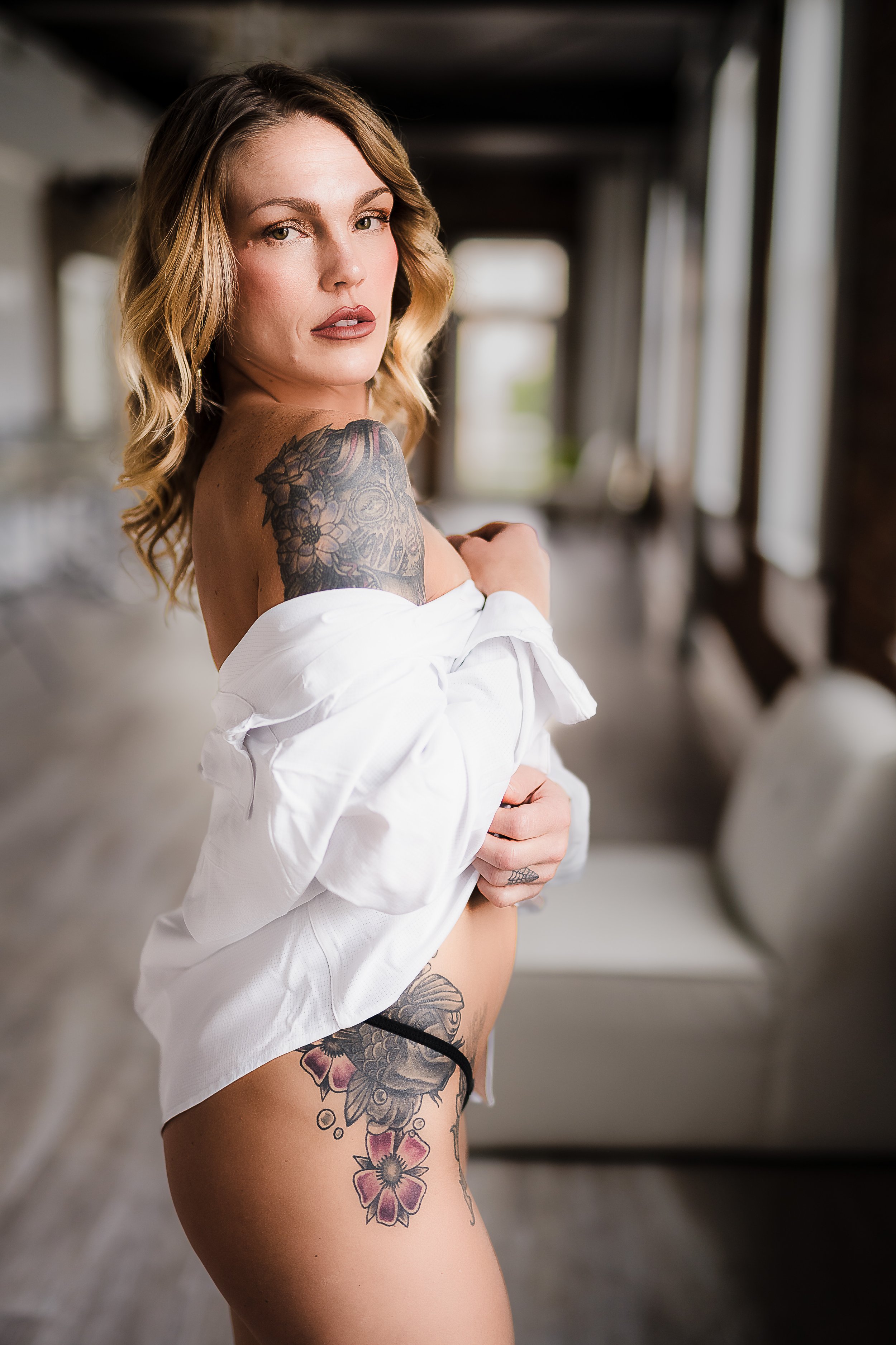 Tattooed woman with a white shirt off shoulder, posing indoors.