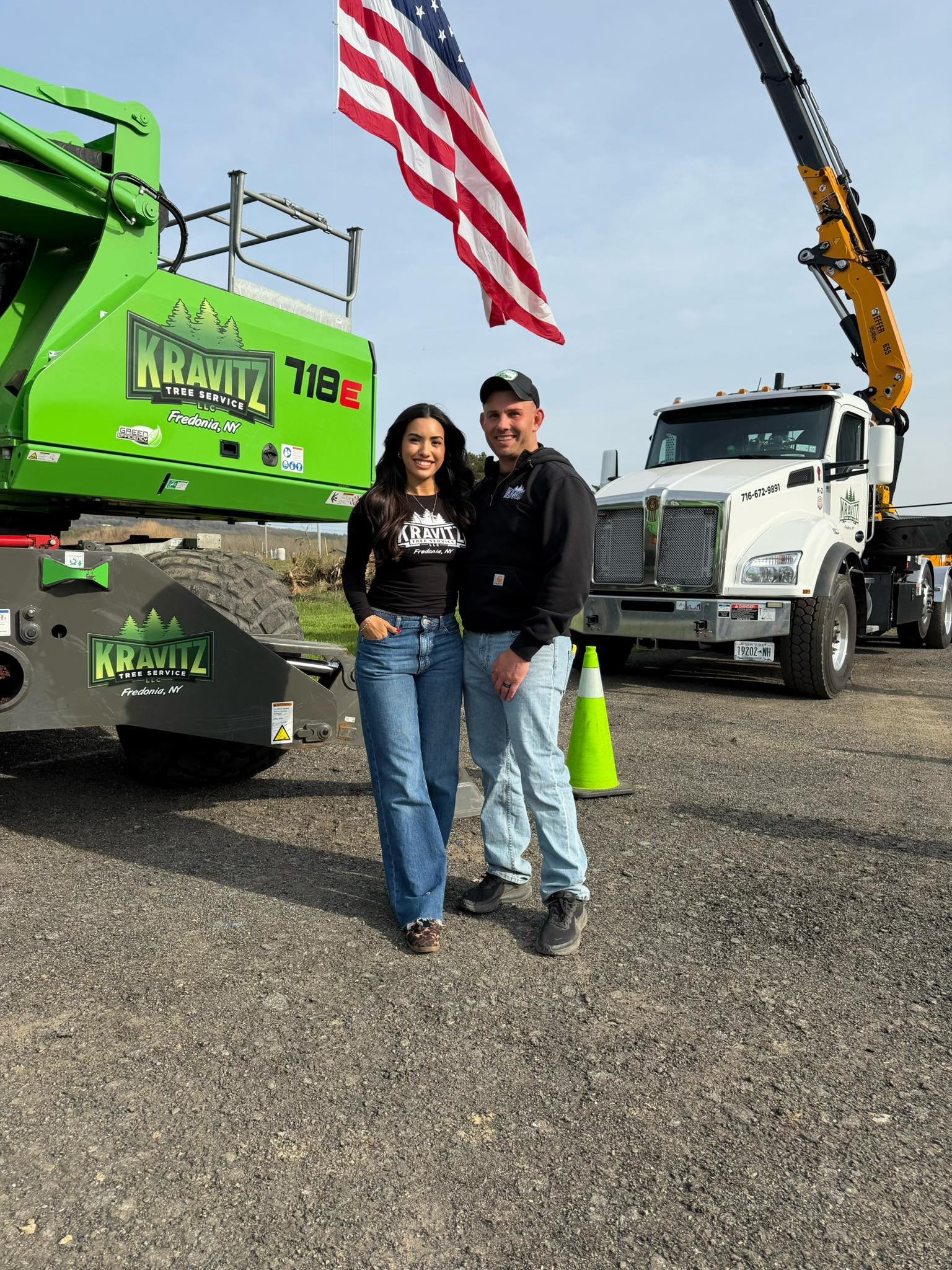 Two people, a woman and a man, standing in front of a large green tree service equipment and a white truck with a crane. The woman is wearing a black Kravitz Tree Service shirt and blue jeans, and the man is wearing a black hoodie and light blue jeans. There is an American flag flying above them and a bright orange safety cone on the ground. The sky is partly cloudy.