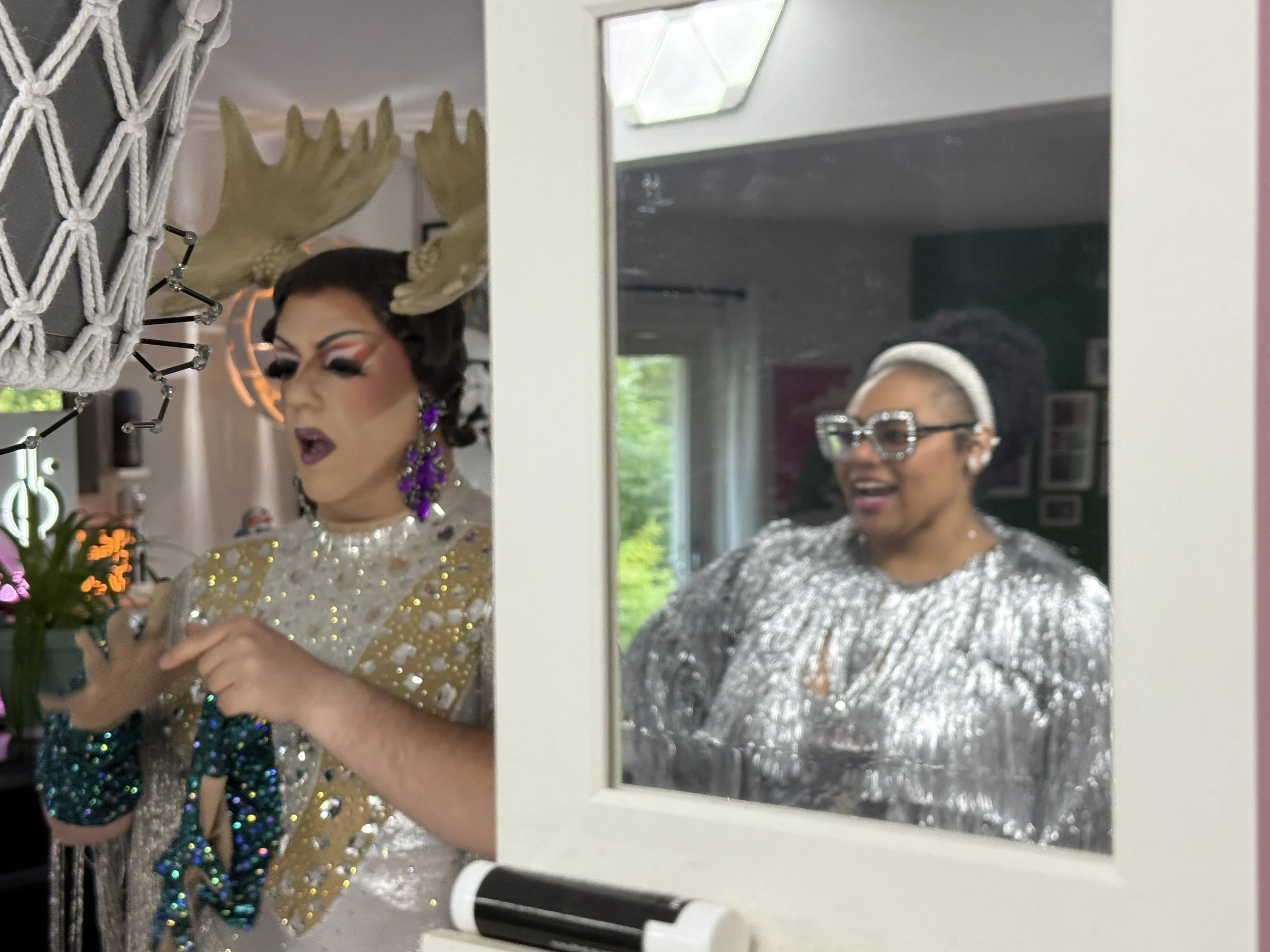 Behind the scenes, Cynthia Mooseknuckle finishes getting into her drag persona as she puts on gloves, while Dr. Amber Nelson talks to her, as seen in a mirror