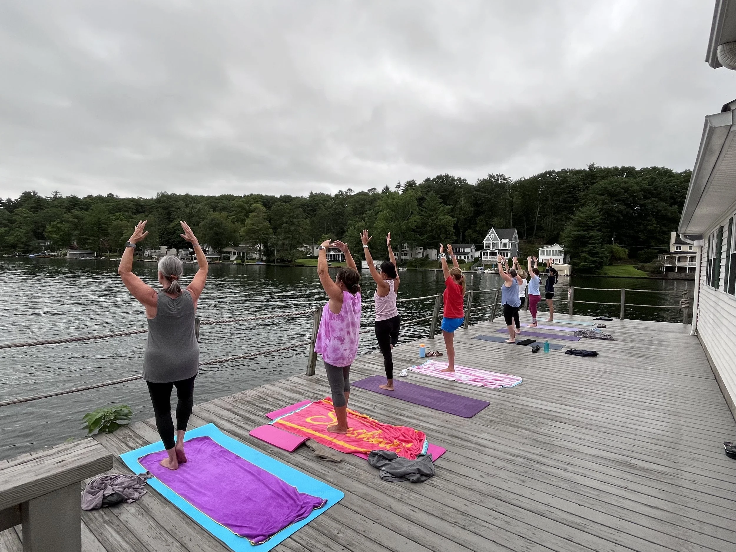 A group of people practicing yoga on a wooden dock by a lake, with houses and trees in the background.