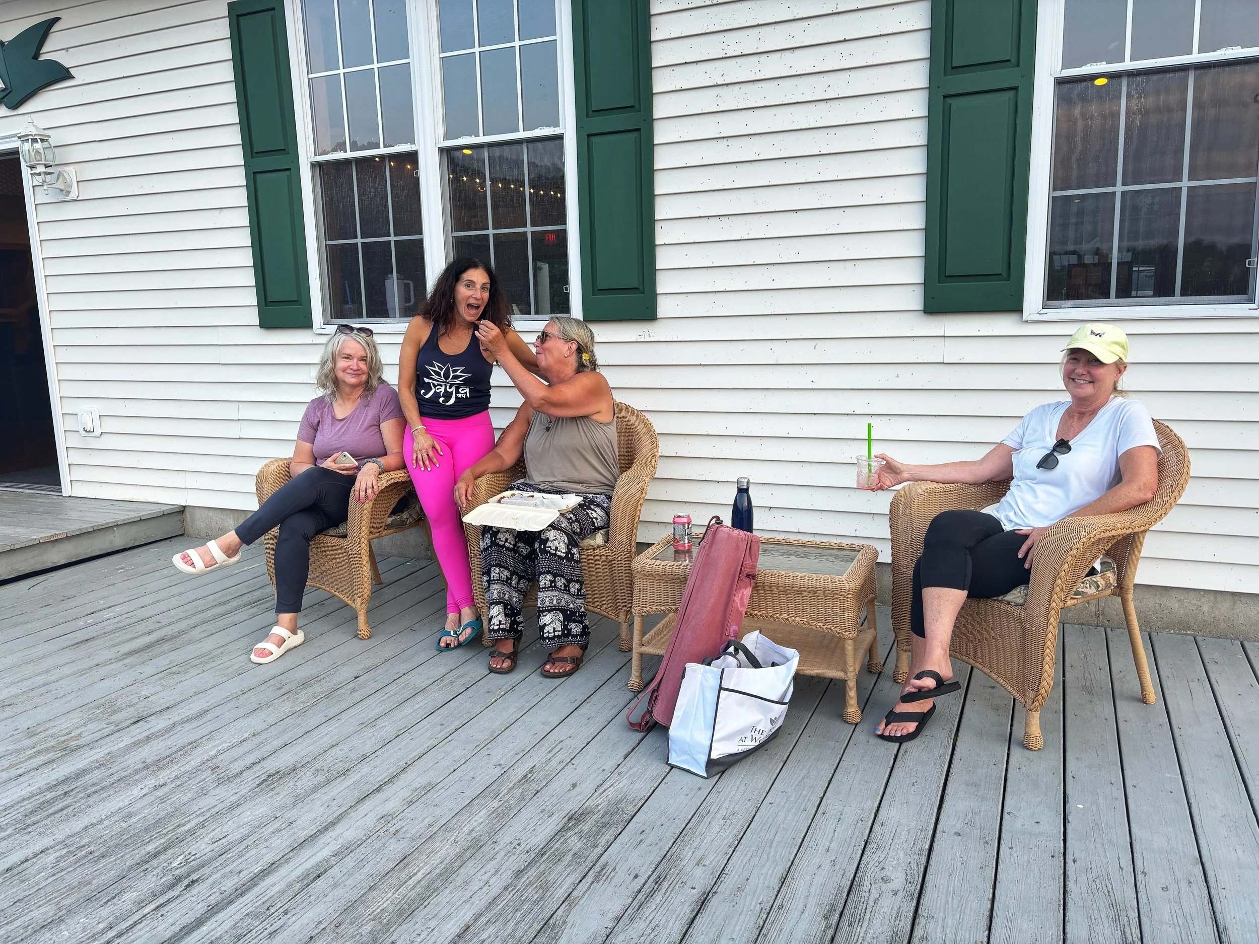Four women sitting and standing on a wooden deck outside a house with white siding and green-shuttered windows. One woman standing in pink pants is being playfully touched on the face by another woman, while two women sit on wicker chairs smiling, on