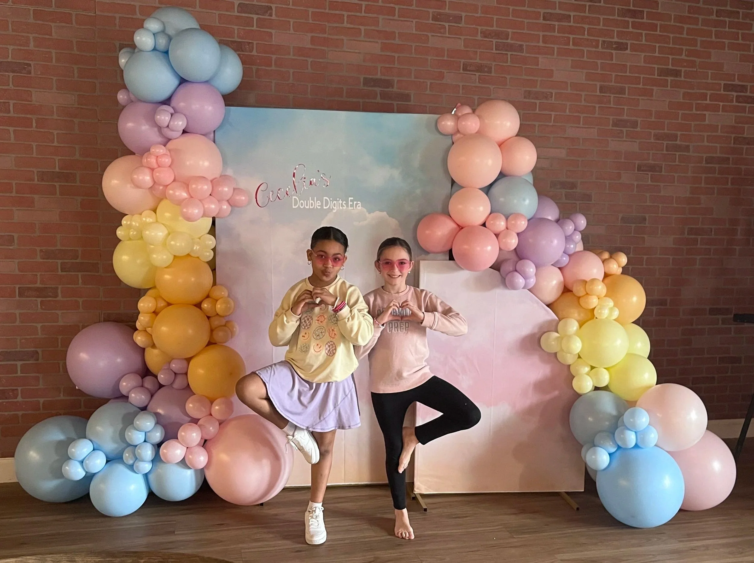 Two girls in front of balloon arch and backdrop, one with yellow sweatshirt and white skirt at a yoga birthday party at Jaya Yoga making heart signs with hands.