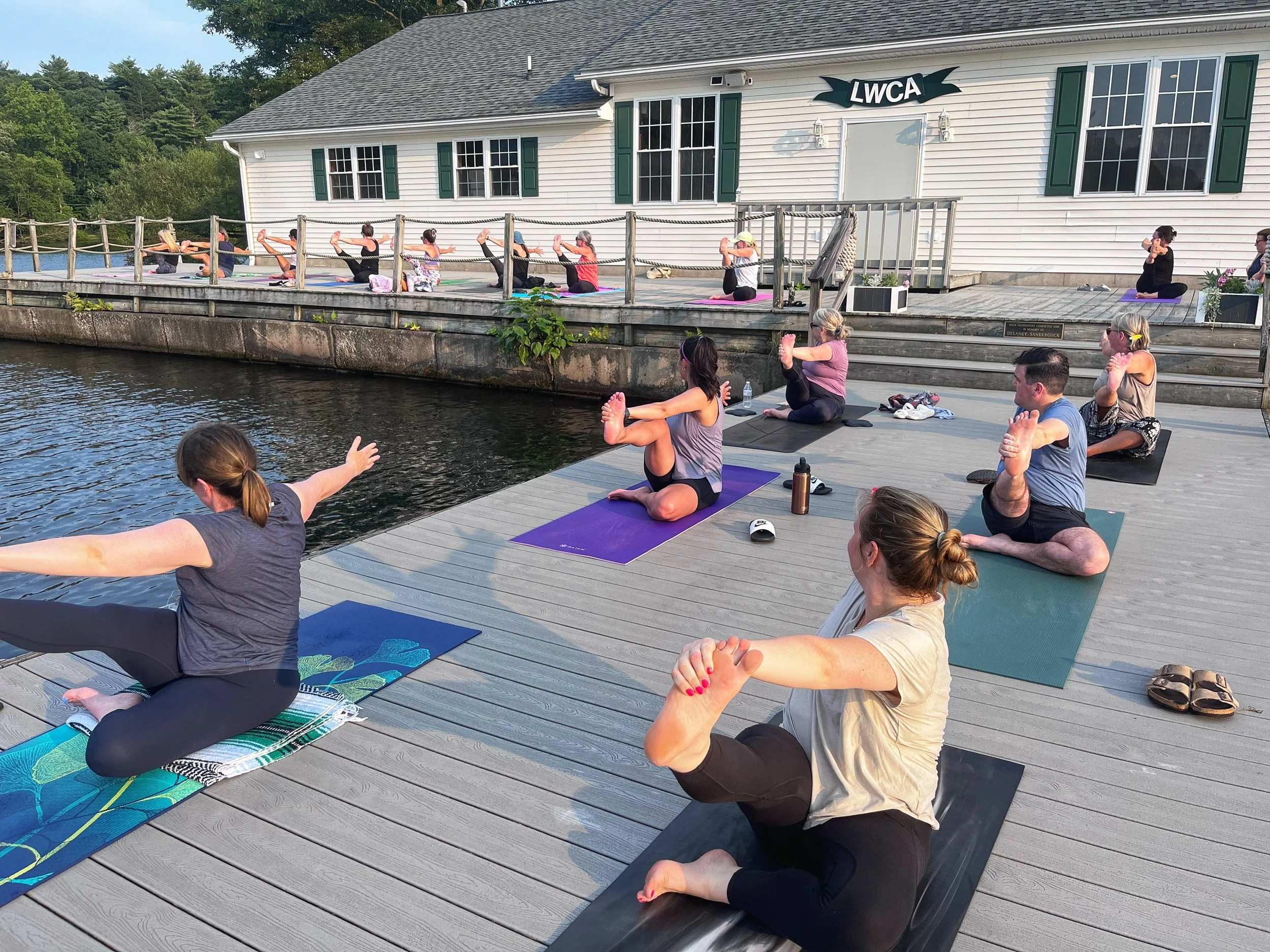 People practicing yoga on a wooden dock by a lake, with an instructor leading the session, in front of a white building with green shutters and a sign that reads "LWCA."