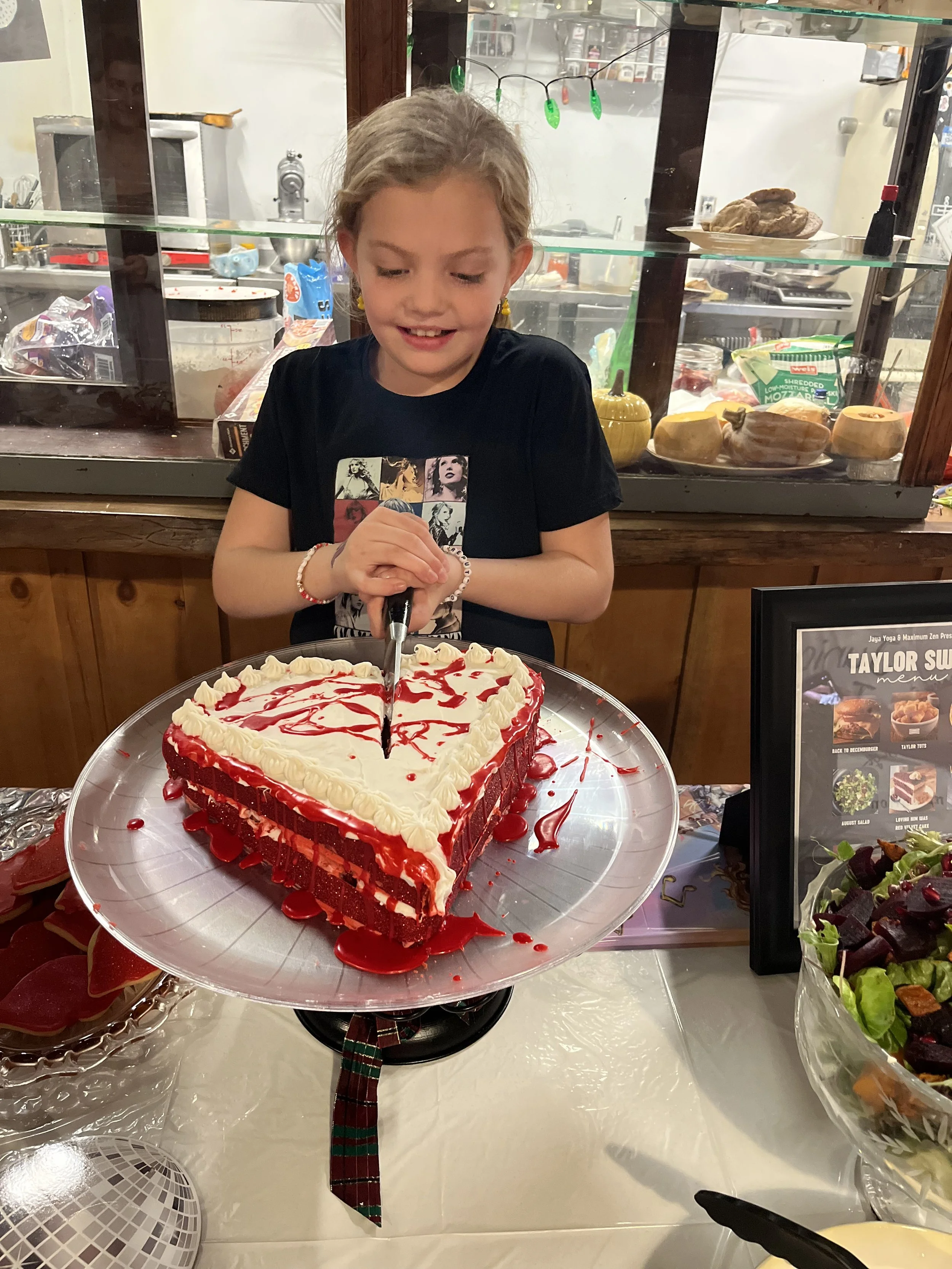 A young girl with a black t-shirt decorated with Taylor Swift images is smiling as she cuts into a heart-shaped red velvet cake with white frosting and red drizzles, decorated with white piped borders at Jaya Yoga Studio.