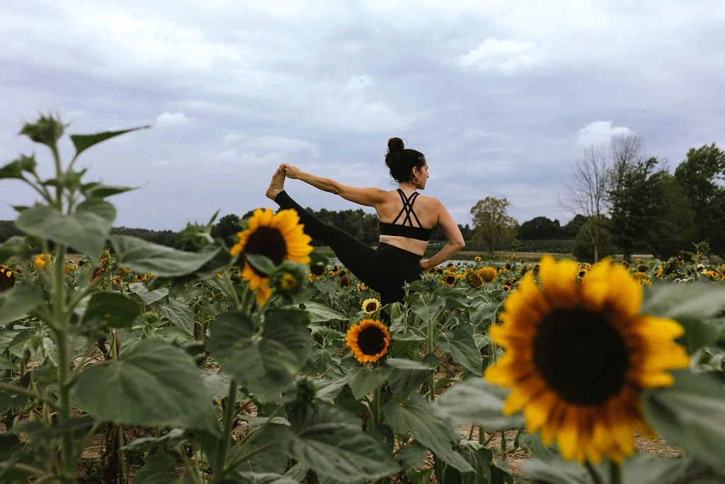 Practicing Hand-to-Big-Toe yoga pose in a sunflower field, demonstrating balance, strength, and mindful practice for every body.