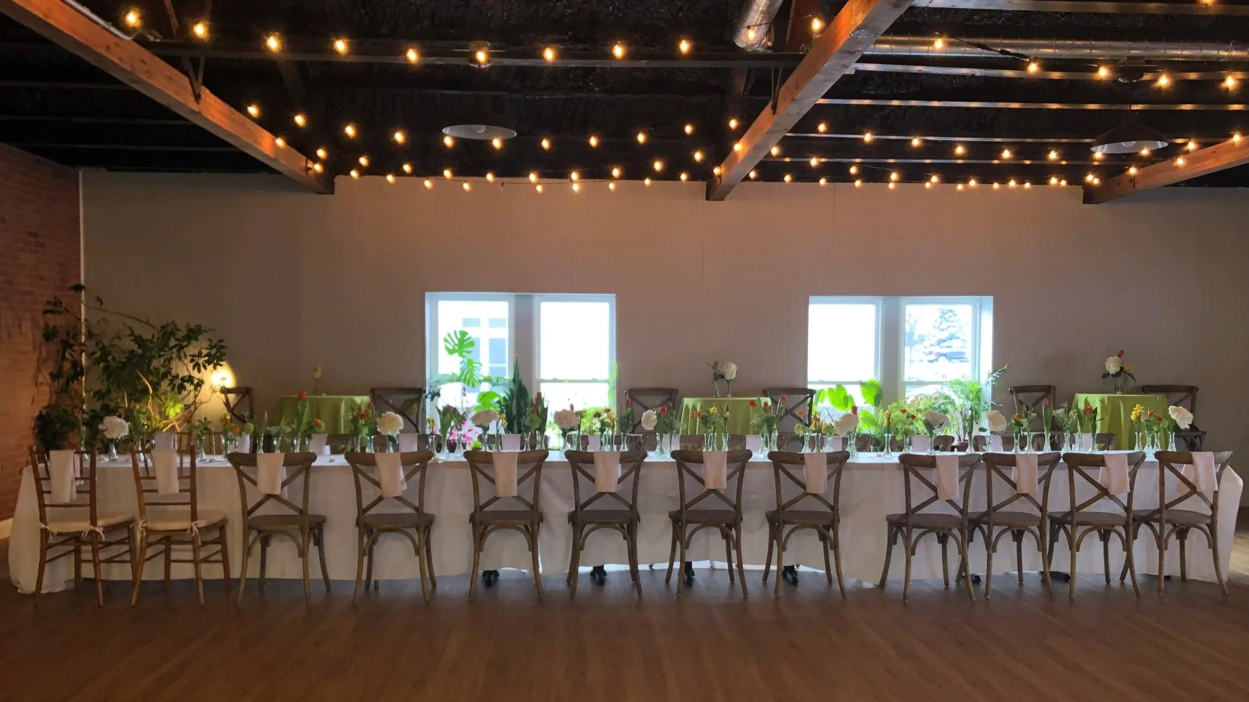 Long banquet table with white tablecloth, decorated with flower arrangements and wine glasses, set up for a celebration in a rustic venue with string lights on the ceiling and large windows revealing greenery outside.