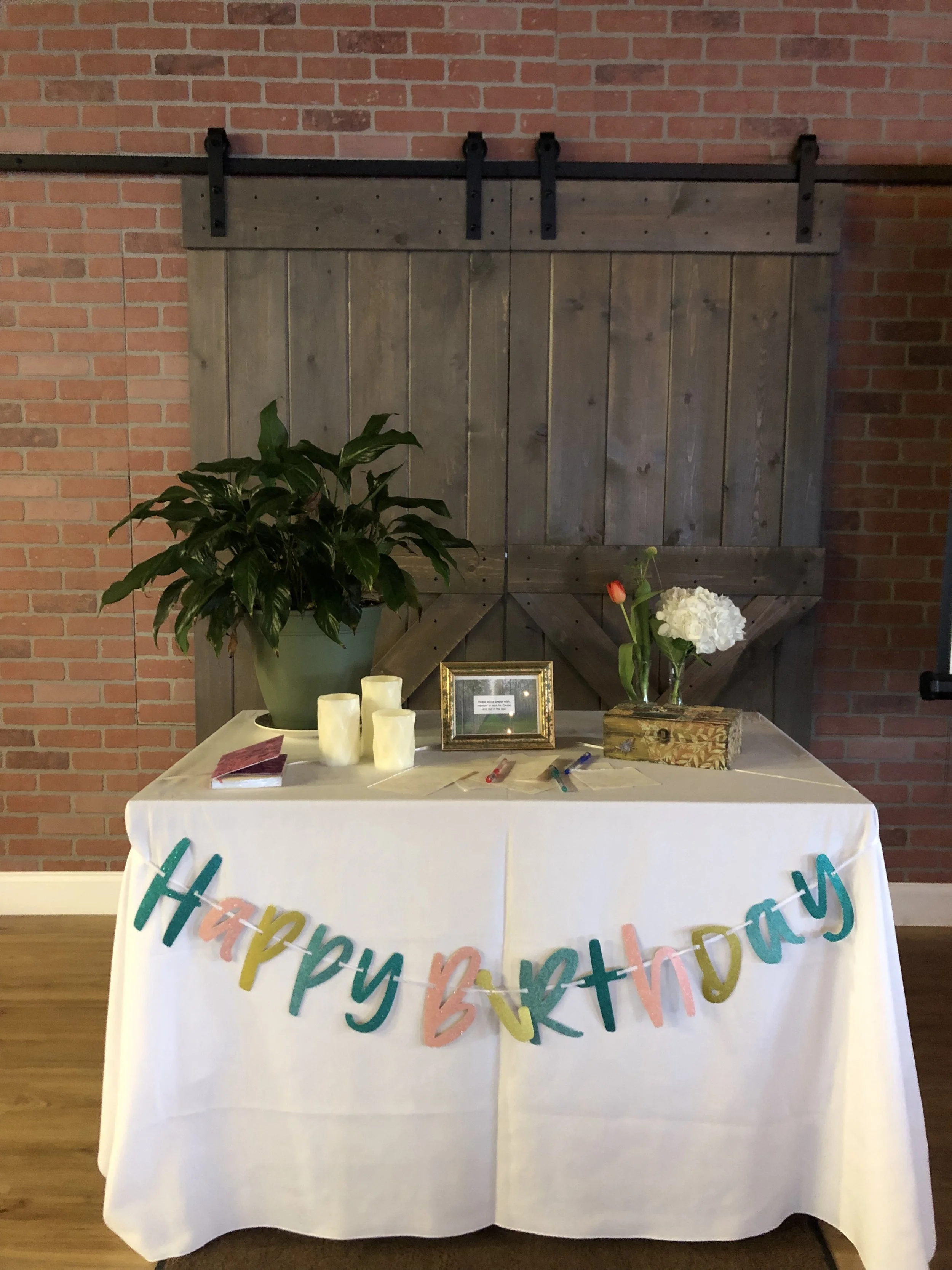 Table with a "Happy Birthday" banner, candles, a potted plant, framed photo, tissue box with flowers, and pens, set against Jaya Yoga Studio's wooden barn door and brick wall.