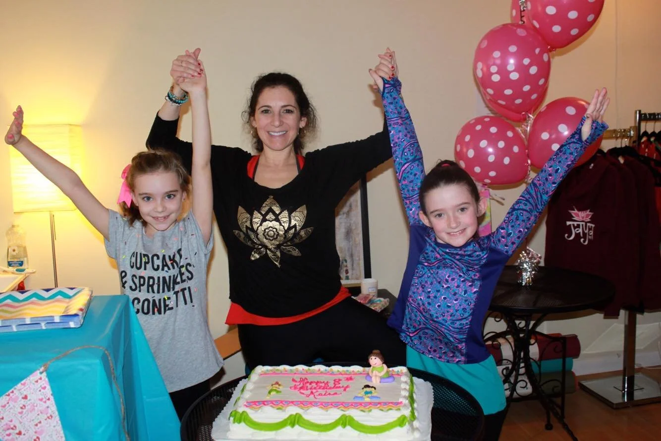 A woman and two young girls celebrating a yoga students birthday with a decorated cake and pink polka dot balloons. The woman and girls are smiling and raising their arms in joy.