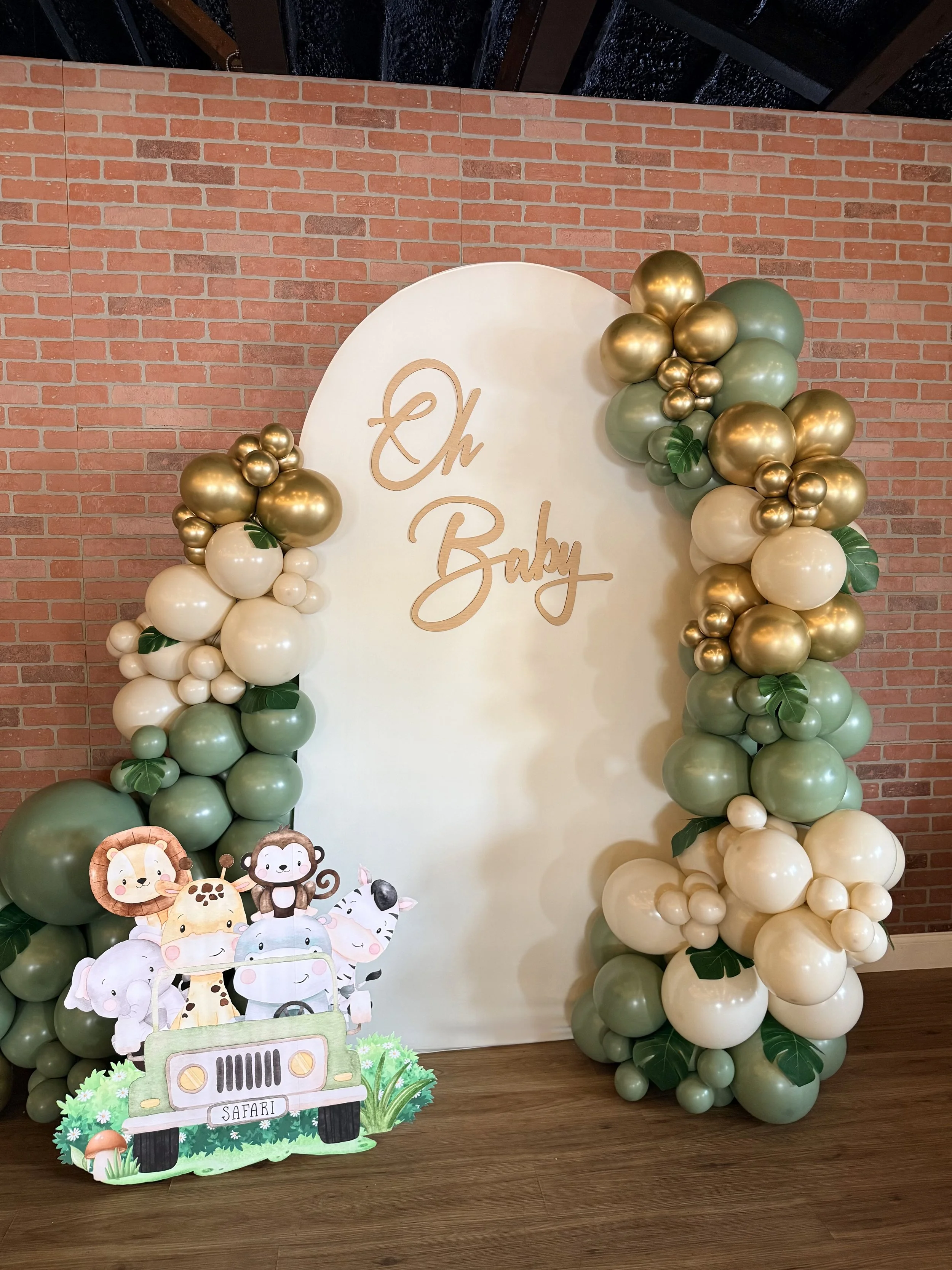Balloon arch with gold, cream, and green balloons surrounding a white backdrop with 'Oh Baby' written in gold script, and a children’s safari-themed paper cutout with cartoon animals in a vehicle labeled 'Safari' in front of a brick wall and wooden f