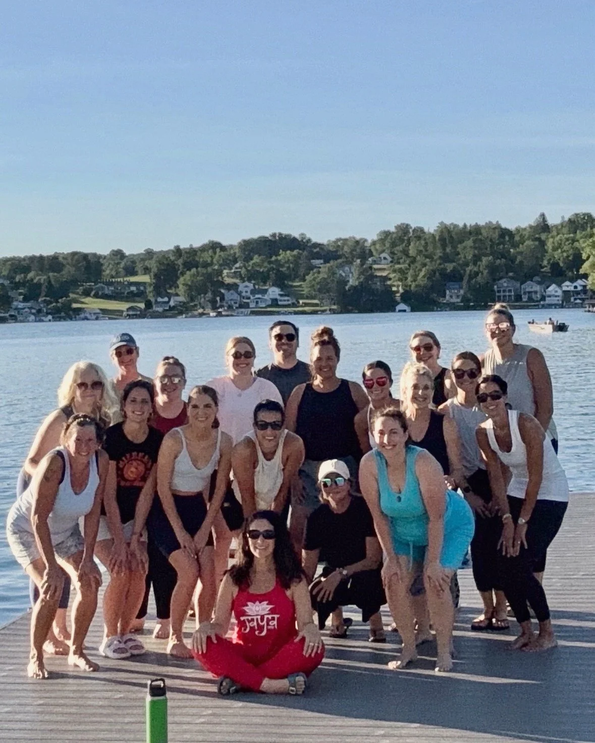 Group of people sitting and standing on a dock by a lake during sunset, with trees and houses in the background after a yoga class on the dock at Lake Winola hosted by Jaya Yoga Studio.
