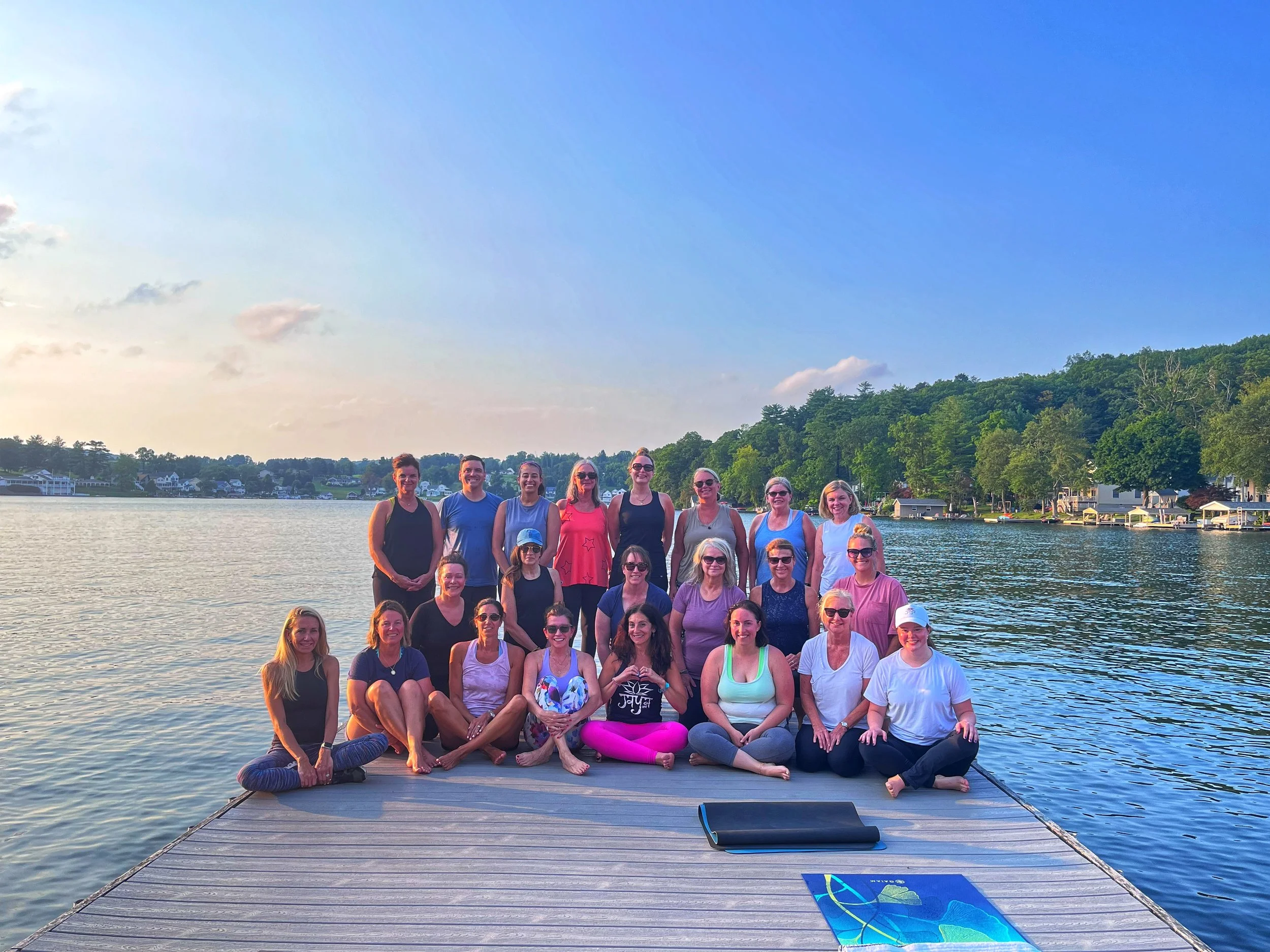 Group of 19 women sitting and standing on a dock by a lake during sunset, with trees and houses in the background.