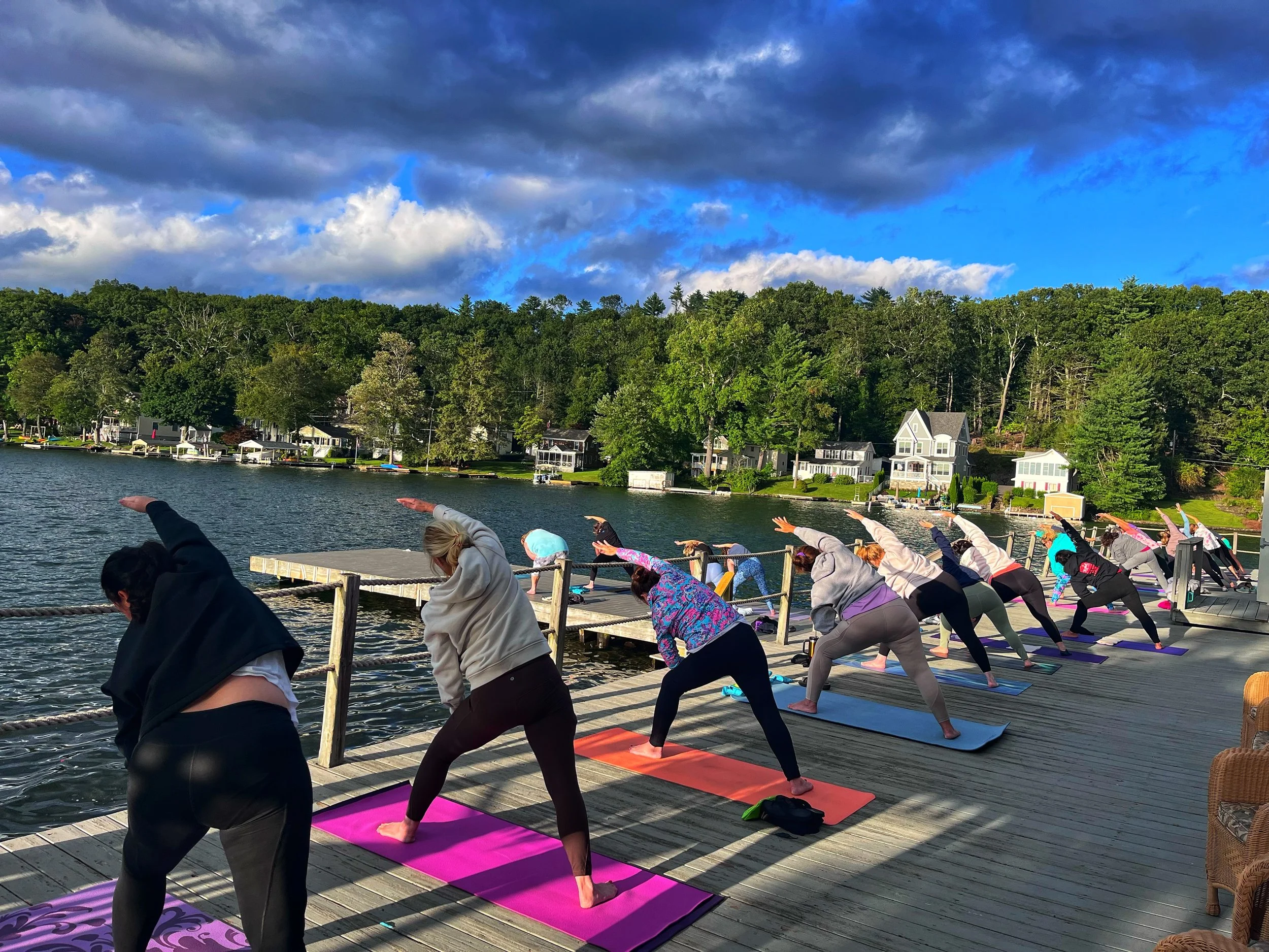 Group people practicing yoga on a wooden dock by a lake during daytime, with a forested hillside and some houses in the background.