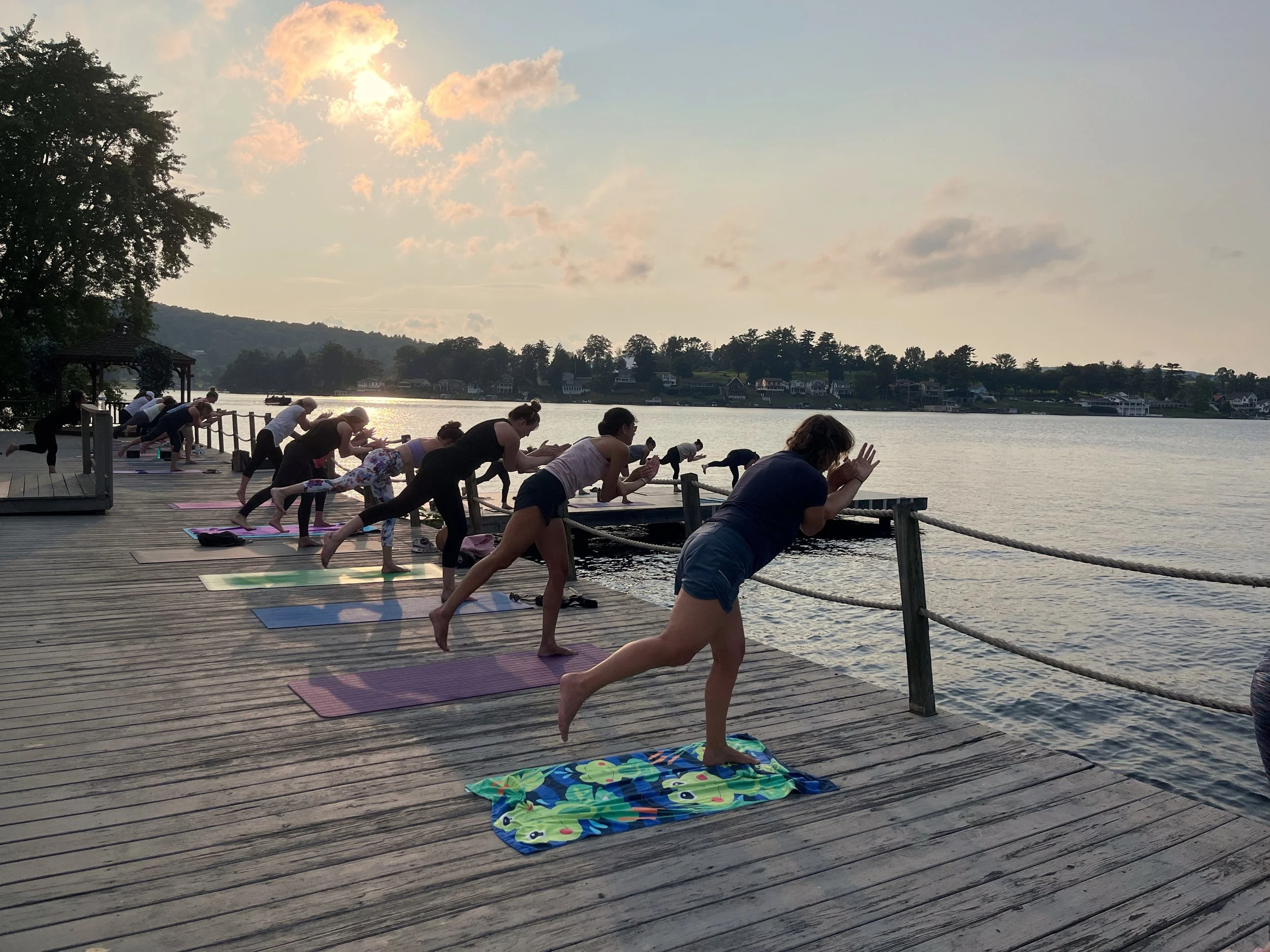 Group of people practicing yoga on a dock by a lake at sunset, all in a balancing pose with one leg raised and hands together.