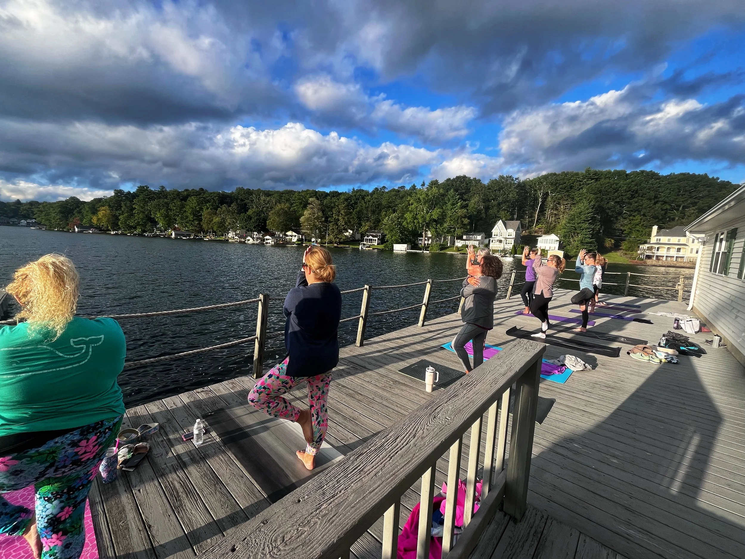 Group of people practicing yoga on a wooden dock by a lake during sunset, with trees and houses in the background and a partly cloudy sky above.