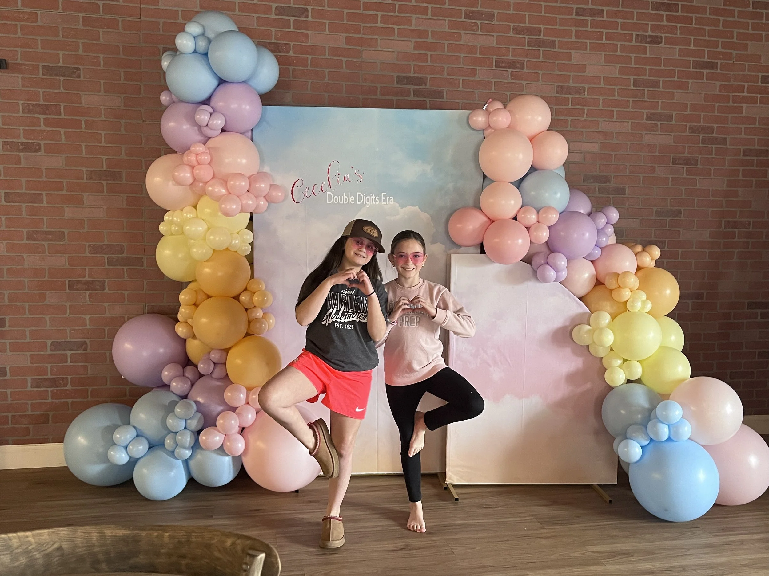Two girls with pink sunglasses making heart signs with their hands, standing on one leg, in front of a rainbow-colored balloon arch and a pastel backdrop with clouds, during a yoga birthday party.