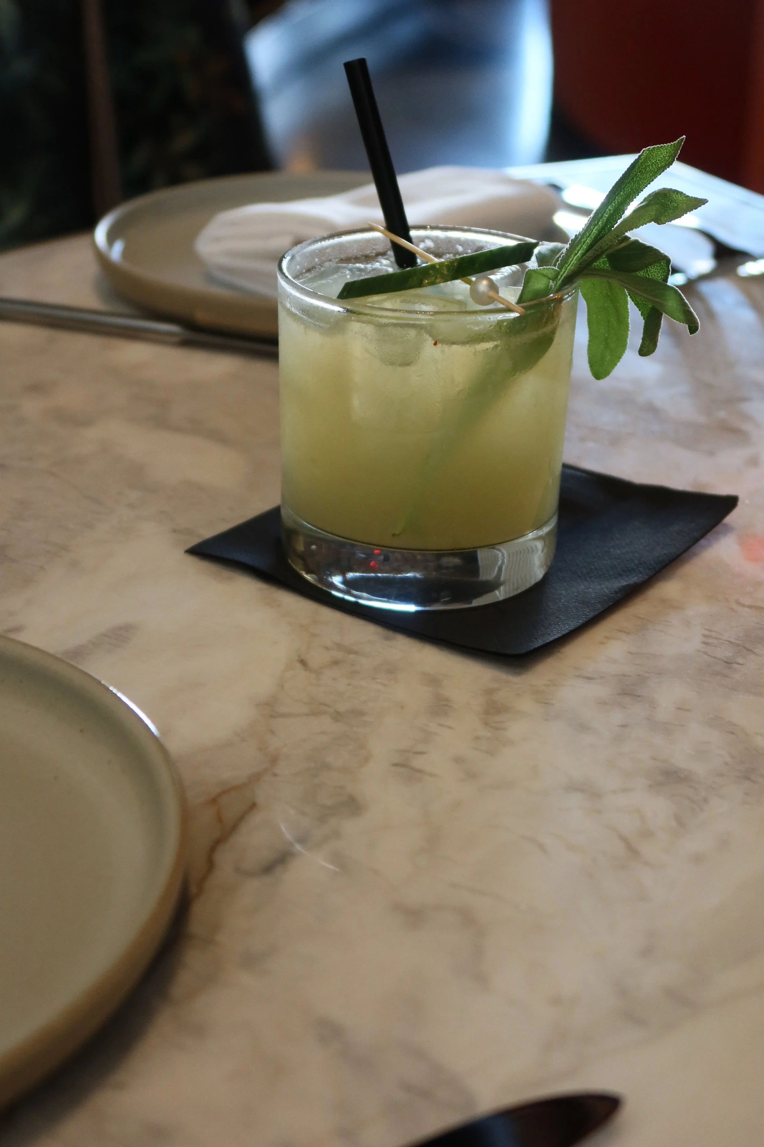 A glass of light yellow cocktail garnished with green leaves and a pearl pin, placed on a black napkin on a marble table.