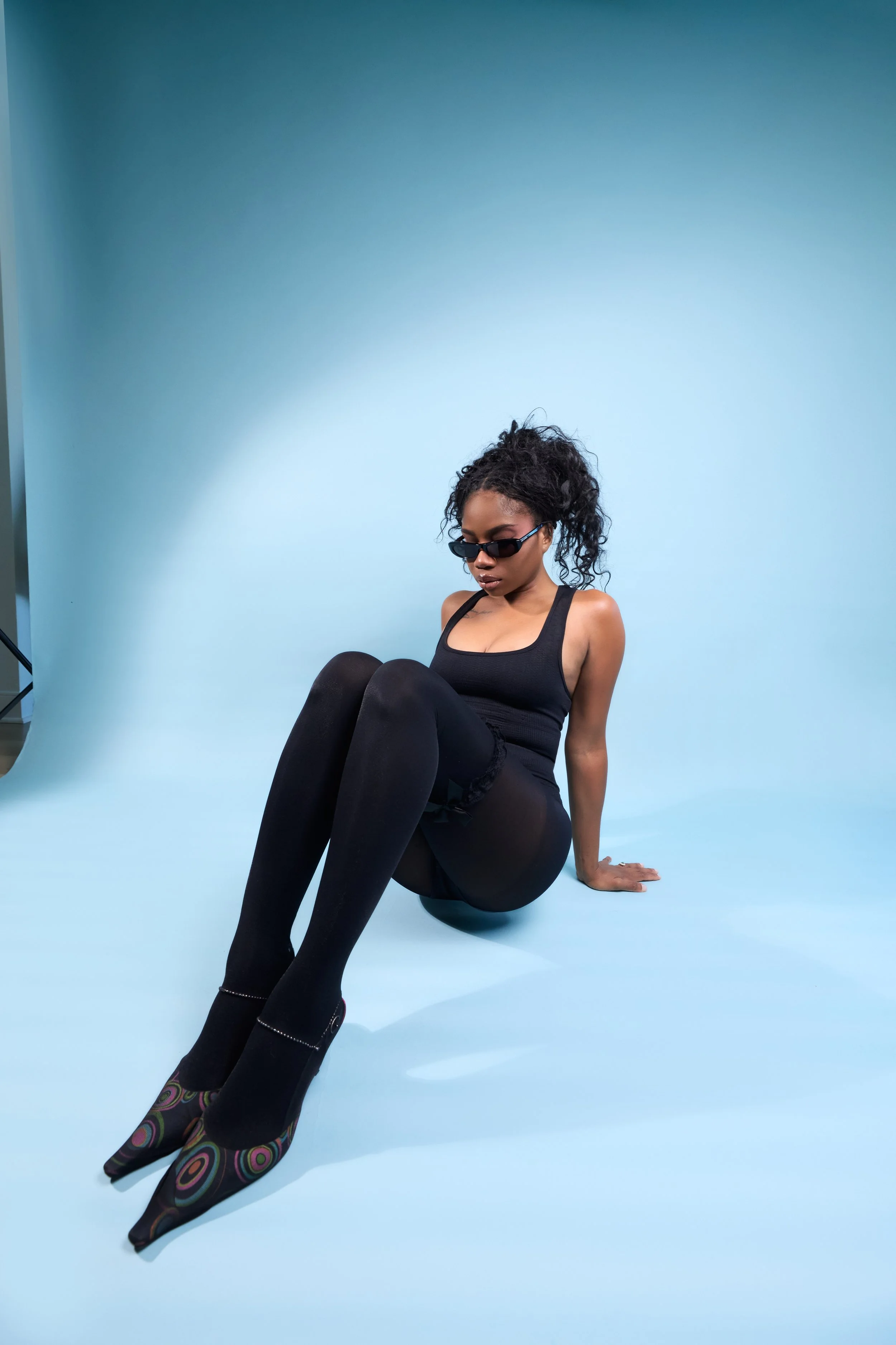 cheyennekimora seated on the ground, arms lifting the body slightly while wearing sunglasses and looking toward knees