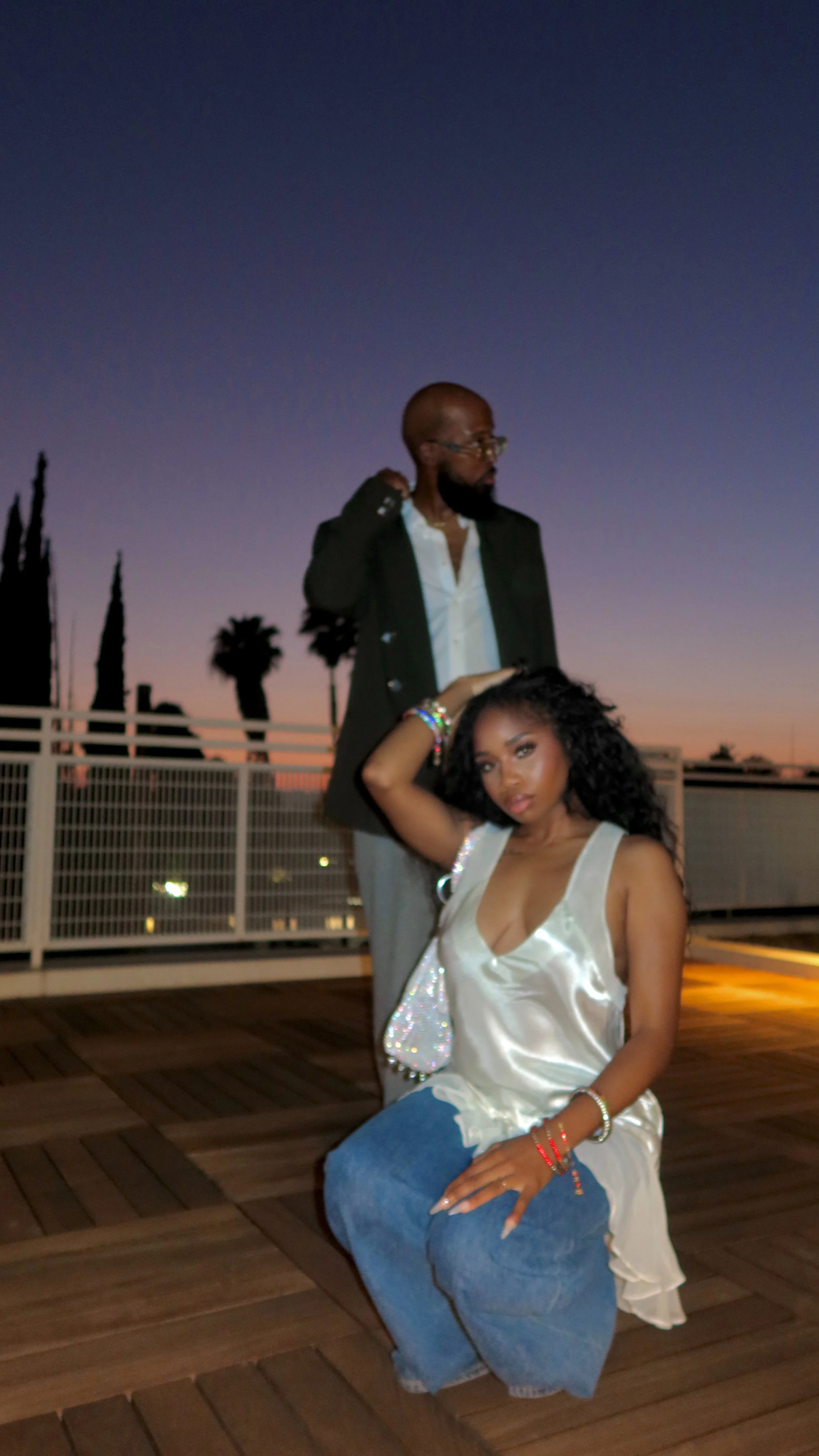 A man standing and a woman sitting outdoors on a wooden deck during sunset, with palm trees and a fence in the background.