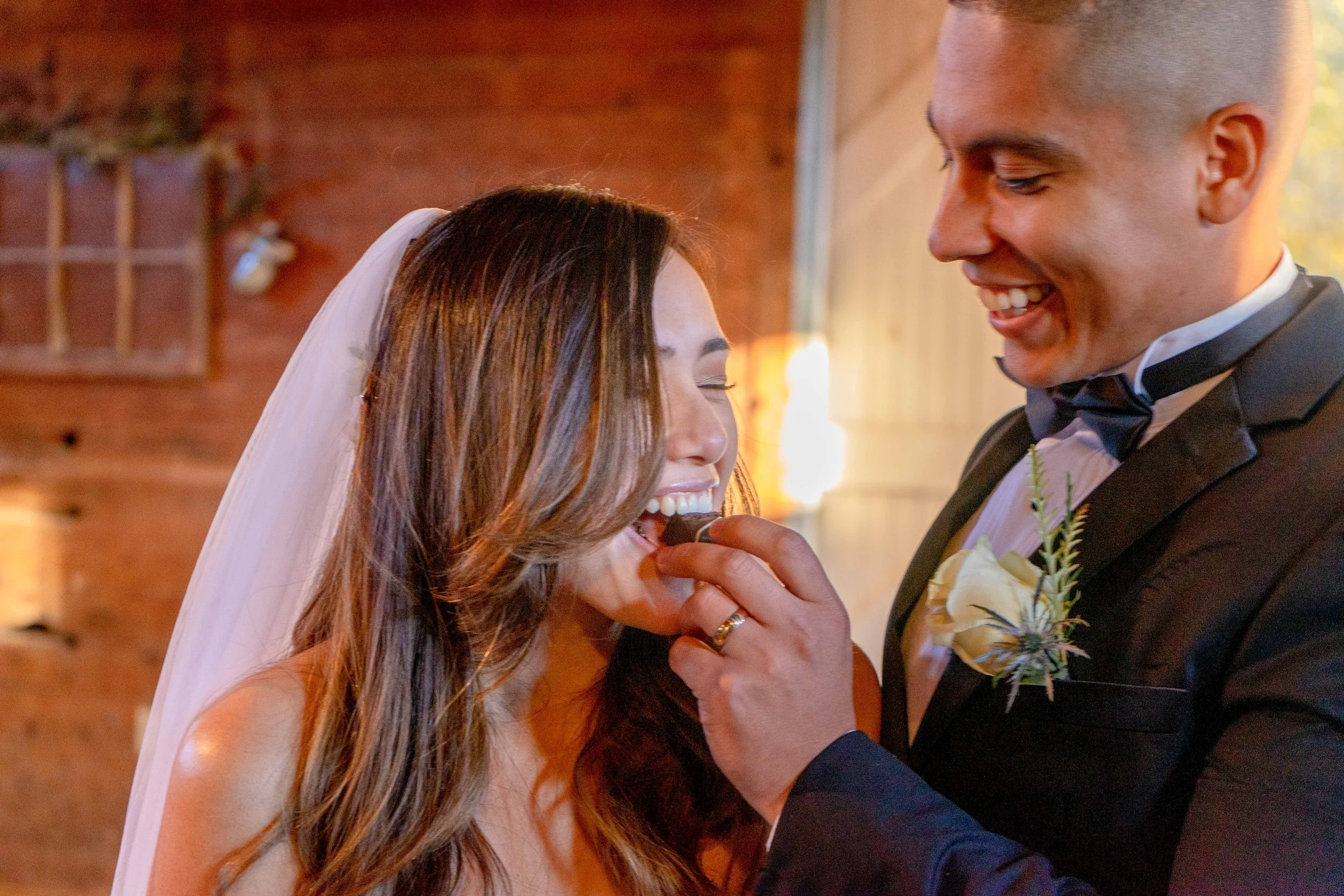 A close-up of a bride and groom during a wedding ceremony, with the groom placing a ring on the bride's finger, smiling and looking at each other, in a warmly lit indoor setting.
