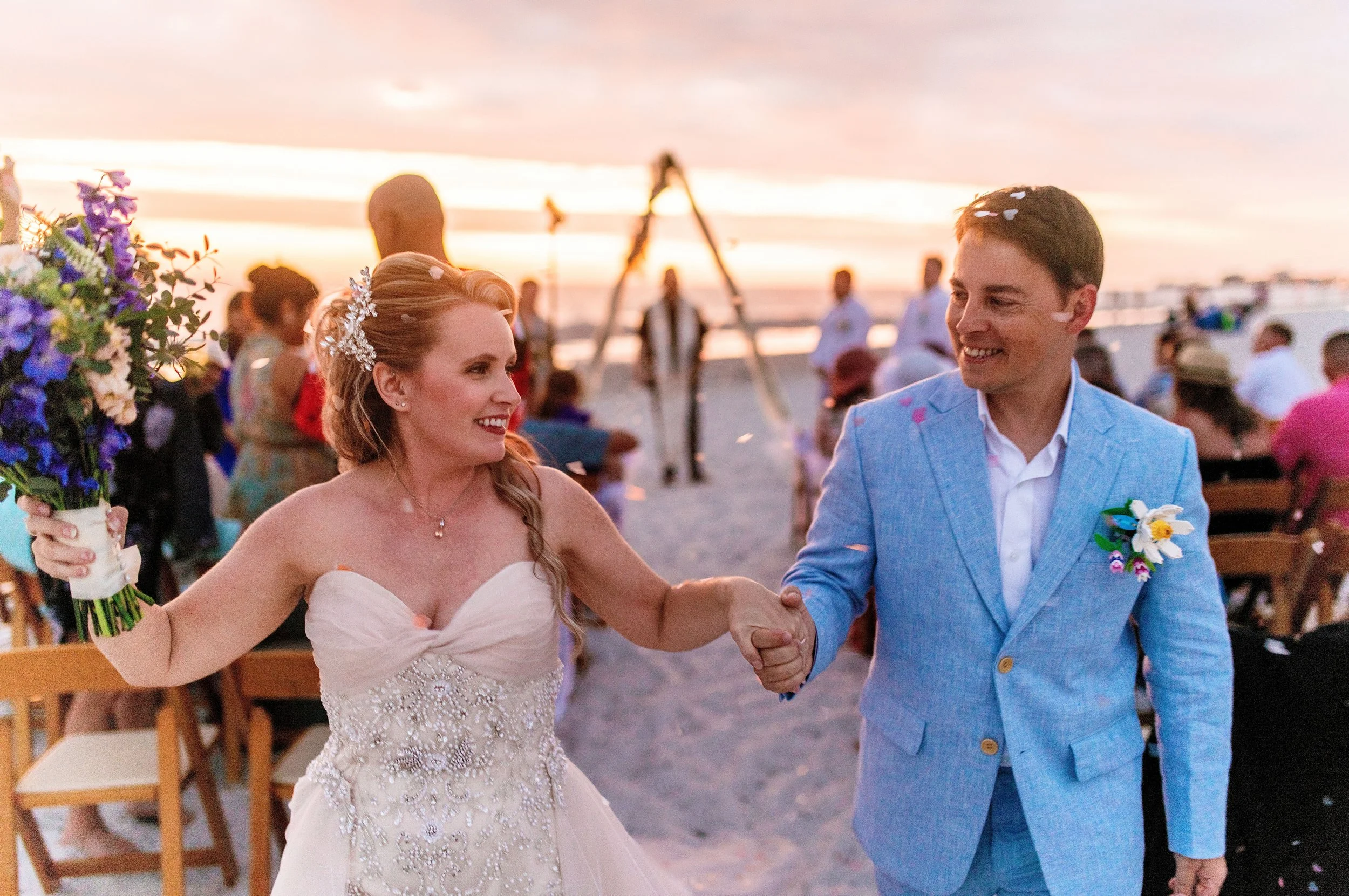 A newlywed couple holding hands and smiling during their wedding on the beach at sunset. The bride is wearing a strapless wedding dress with beadwork, and the groom is in a light blue suit. There are guests and wedding decorations in the background, with a sunset sky and ocean.