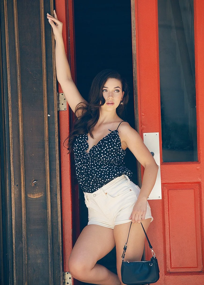 A young woman with dark wavy hair is standing in a doorway, wearing a black polka dot top and white shorts, holding a small black purse, with one arm resting on the doorframe.