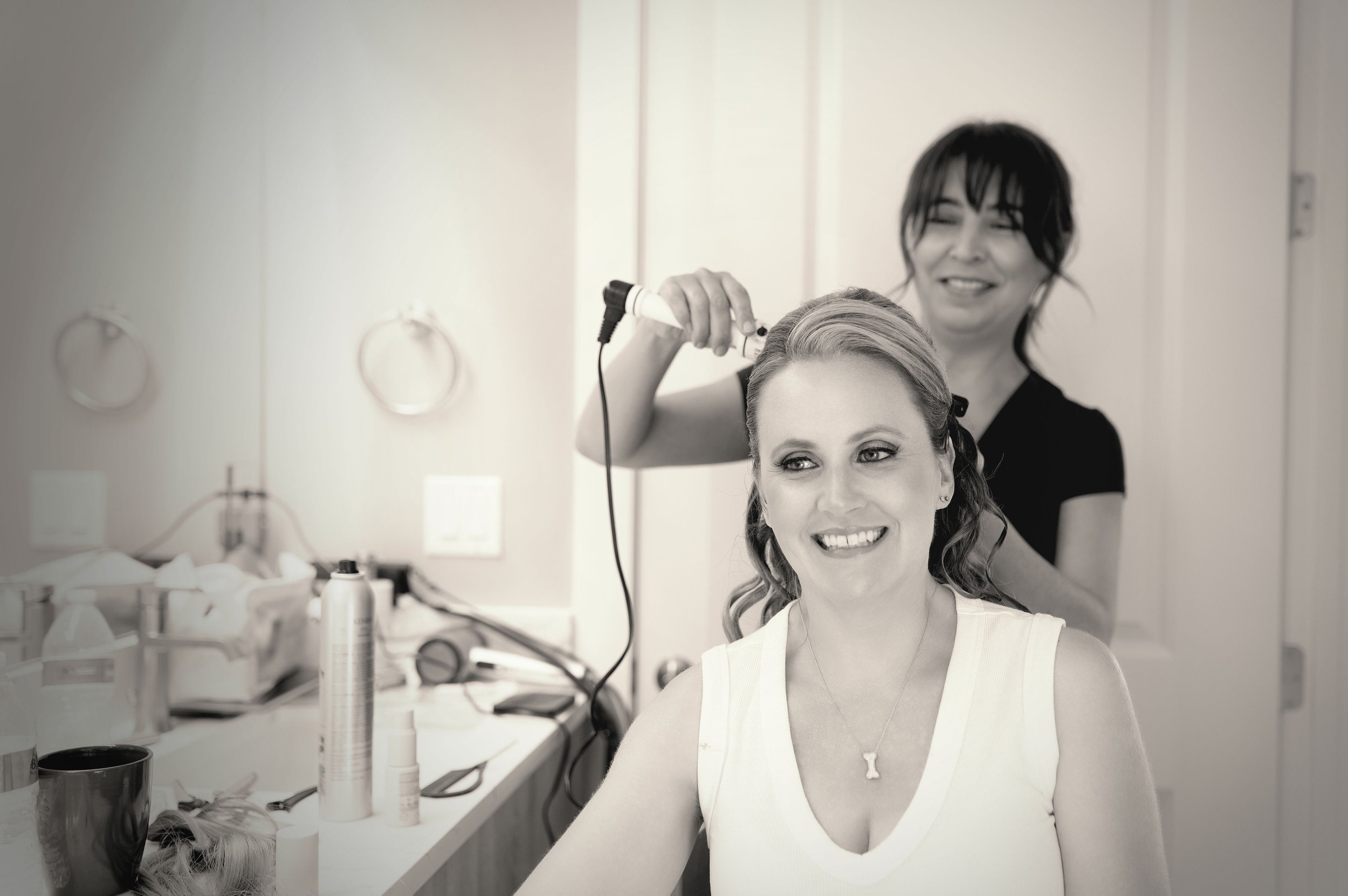 A woman with light-colored hair sitting in a salon chair smiling while a stylist with dark hair styles her hair with a curling iron. Salon tools and supplies are on the counter in front of them.