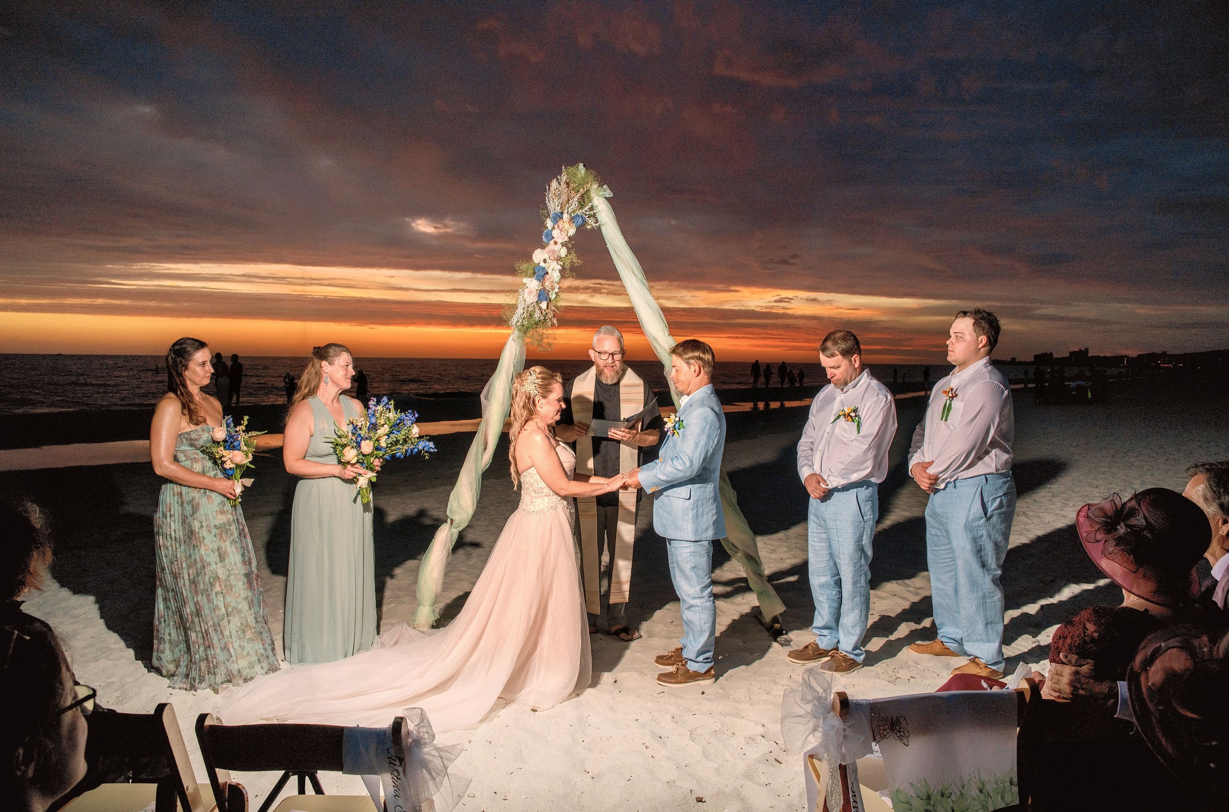 A beach wedding ceremony taking place at sunset with an ocean and cloudy sky in the background. The bride and groom are holding hands under a decorated arch with flowers and fabric. Bridesmaids and groomsmen stand beside them, with guests seated in t